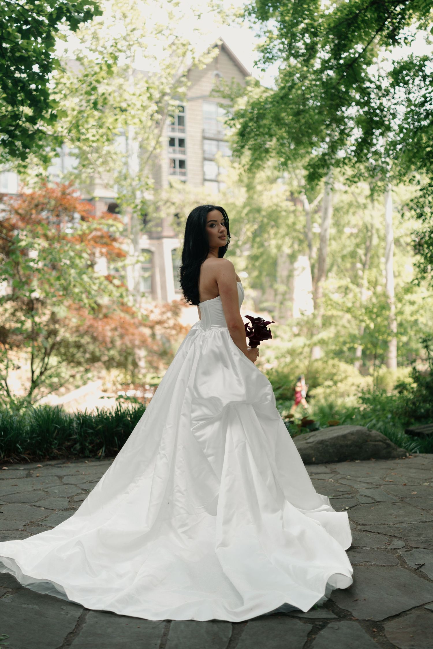 Bride in strapless white ballgown with long train poses in lush garden setting near stone building.