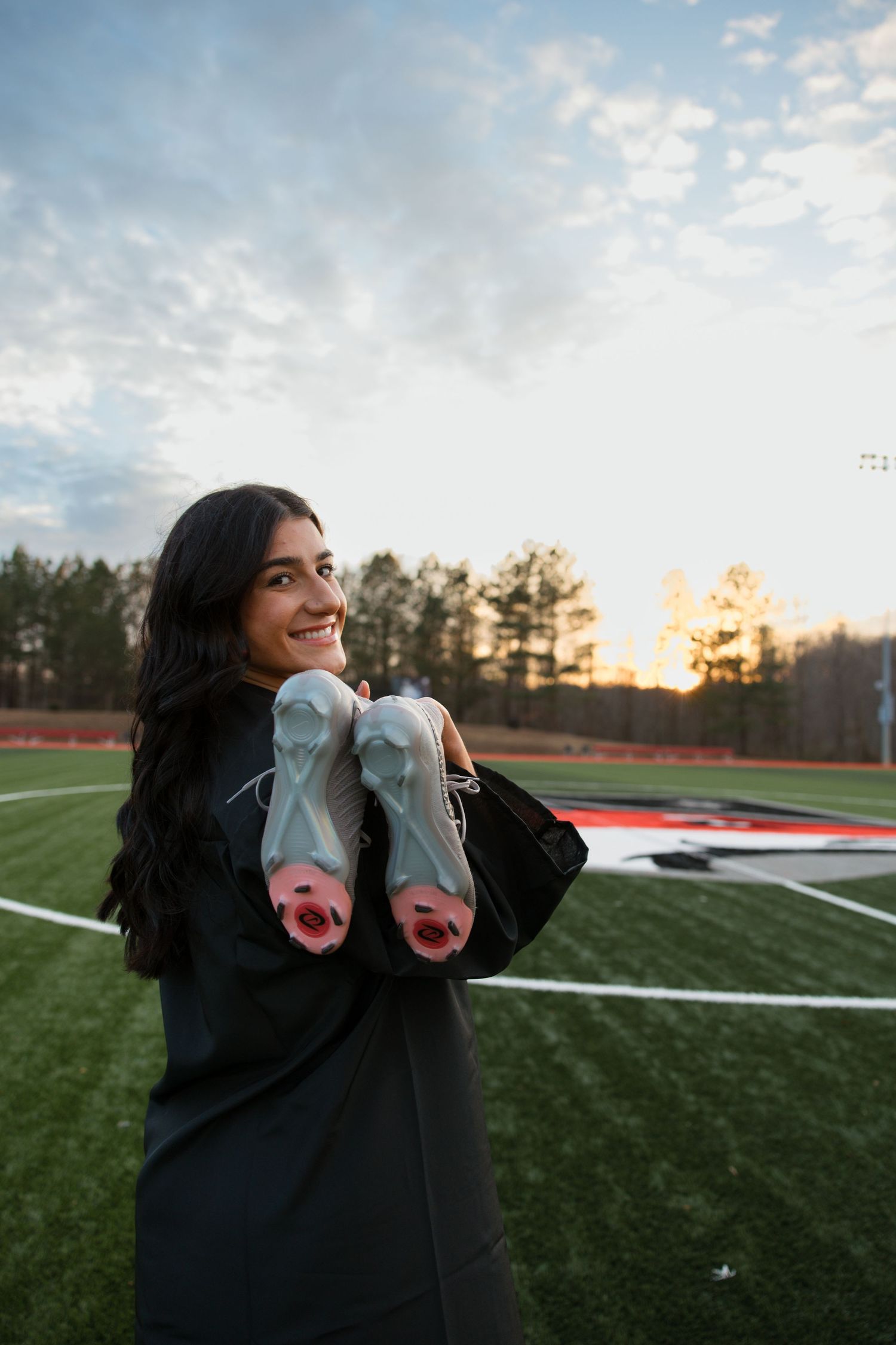 Graduate in cap and gown holds up graduation shoes on sports field at sunset.