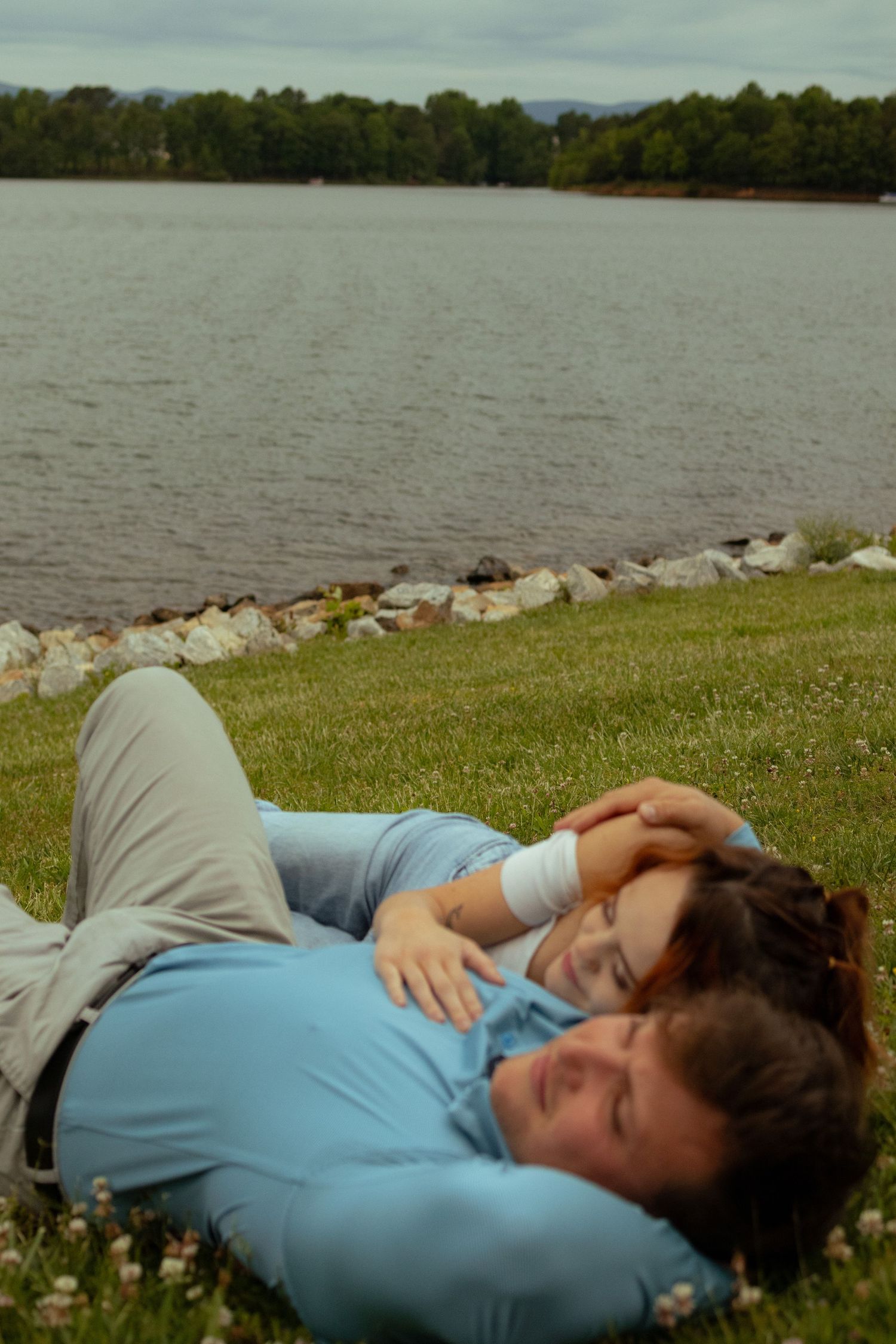 A person in a blue shirt lies peacefully on grassy ground beside a lake with trees visible in the background.