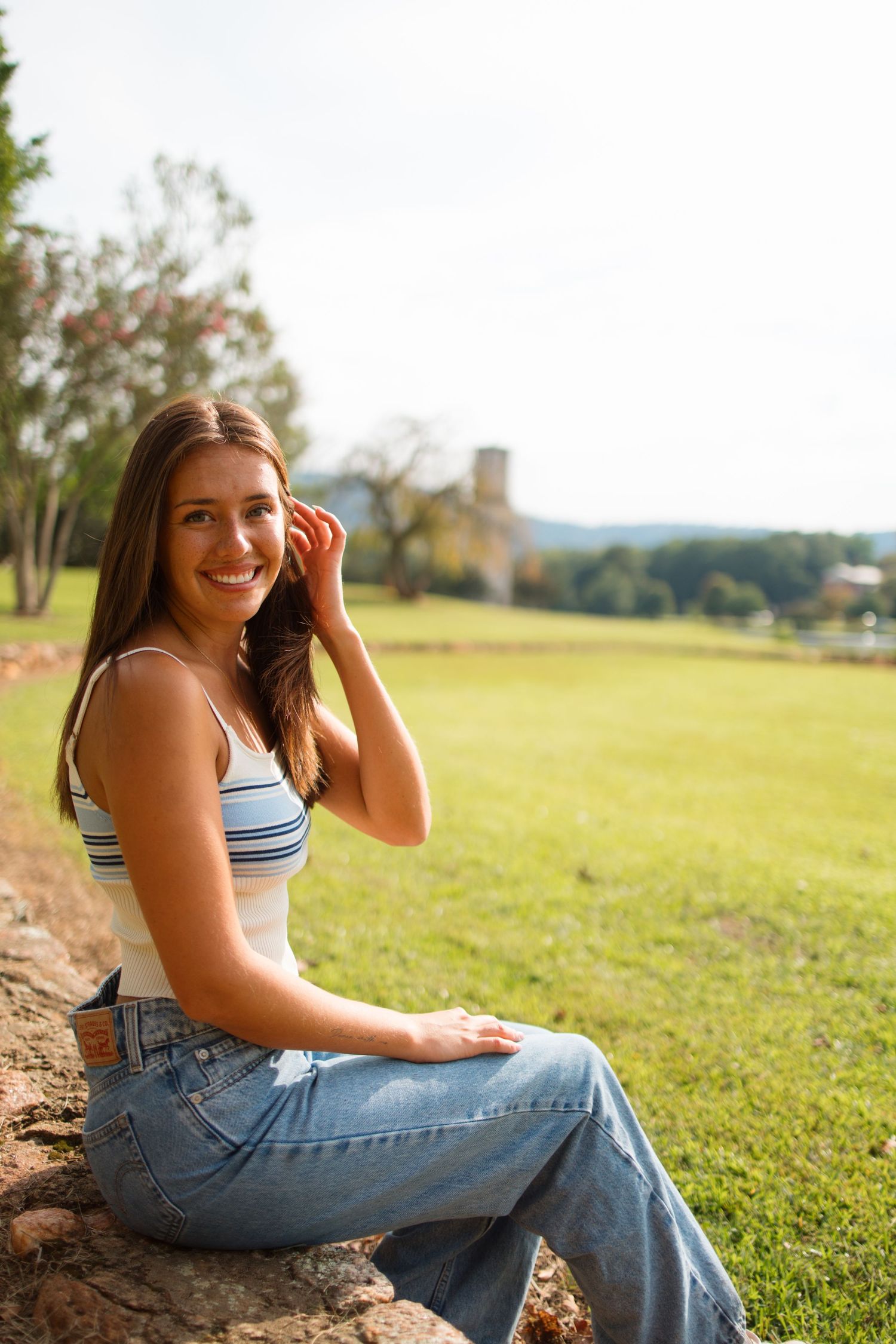 Person in striped tank top and jeans sits on rock wall overlooking sunny grassy field on summer day