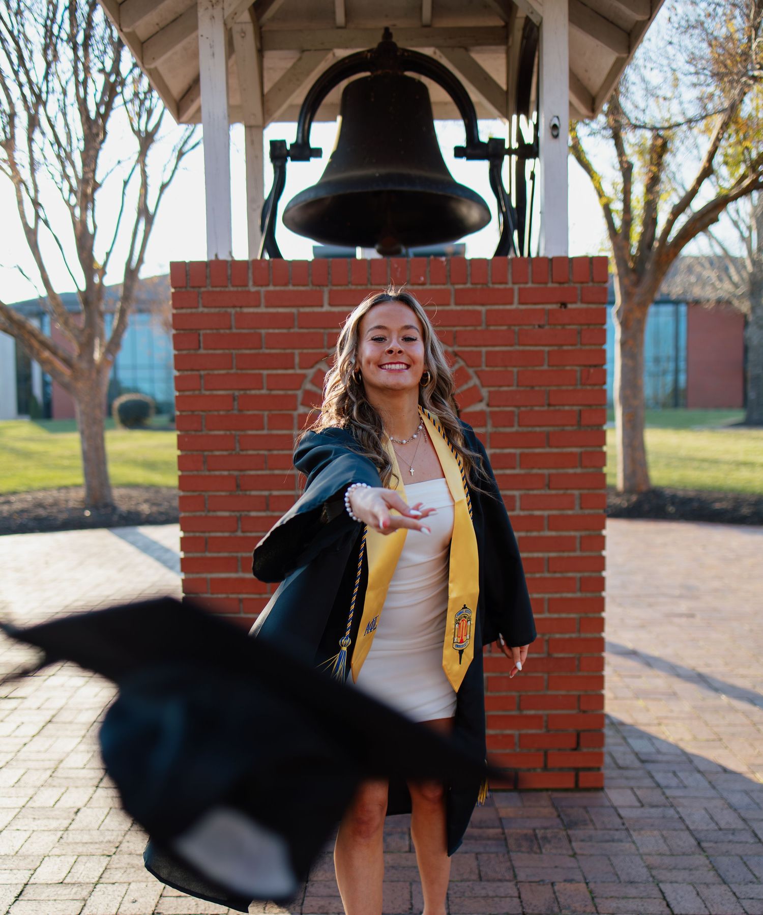 Graduate in academic regalia poses joyfully in front of a brick bell tower.