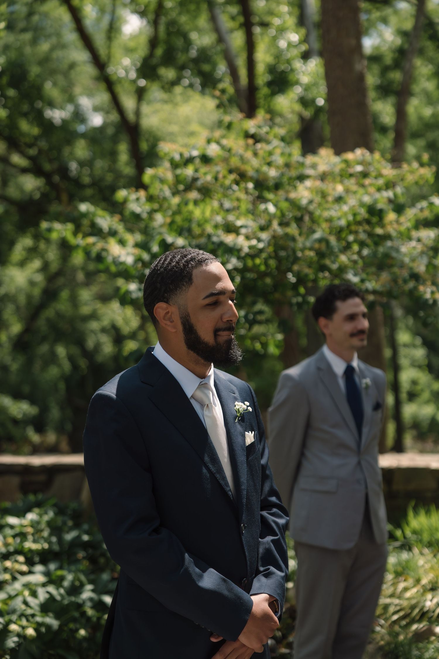 Groom in navy suit with boutonniere stands outdoors at wedding ceremony, groomsman in gray suit visible in background.