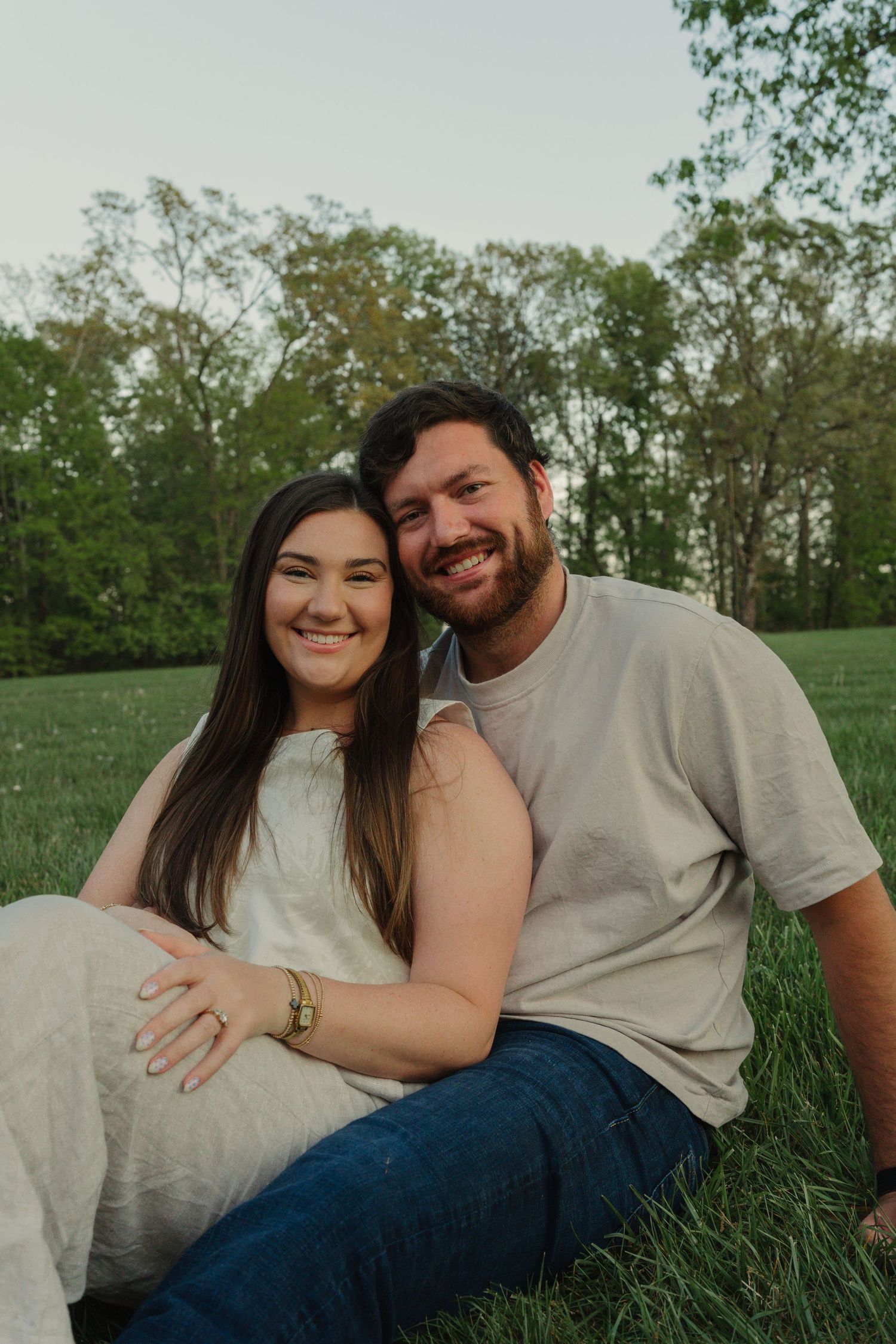 A smiling couple sits together on green grass with trees in the background during golden hour.