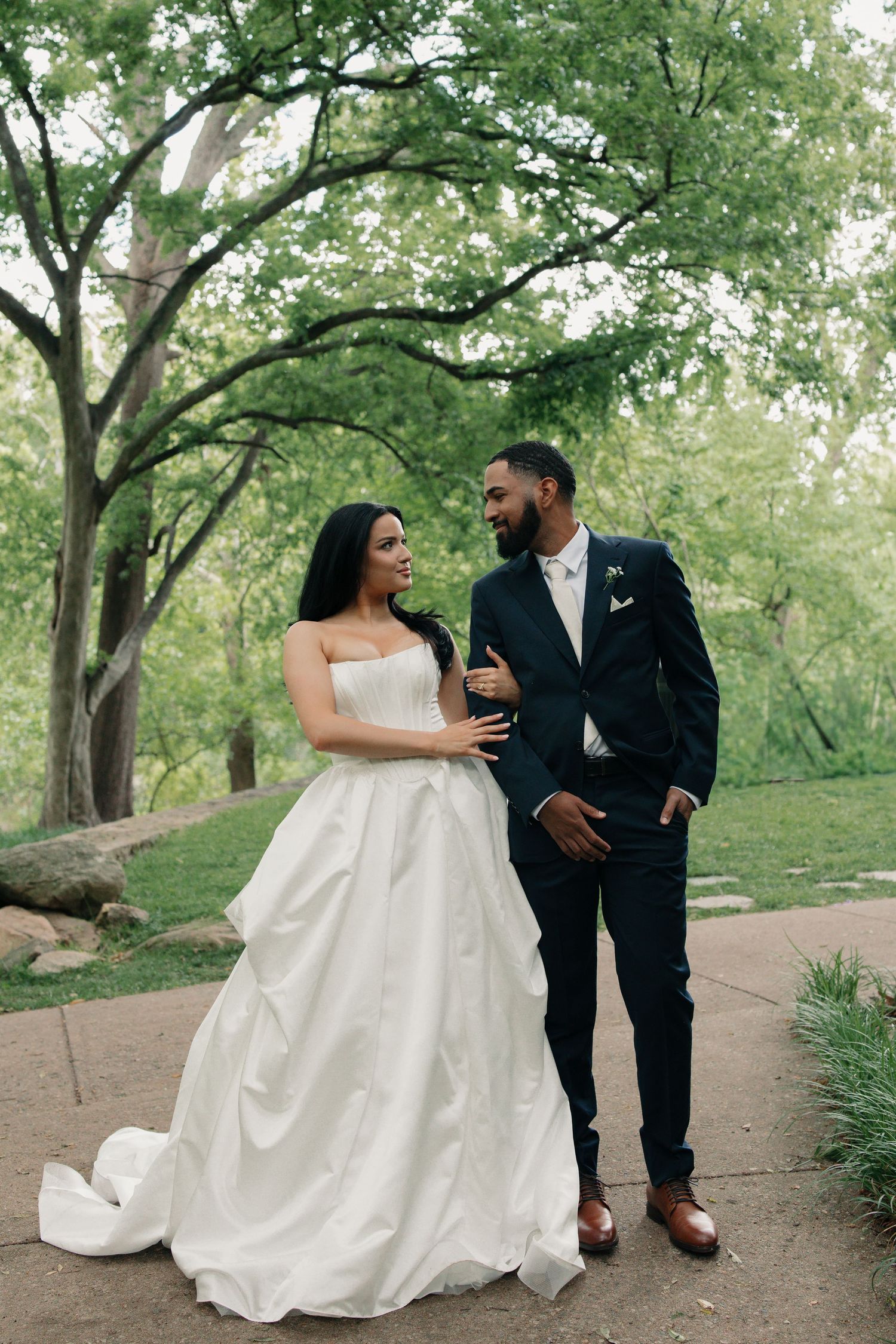 A bride and groom share a romantic moment outdoors, surrounded by lush green trees, as she holds his arm in her elegant white gown.