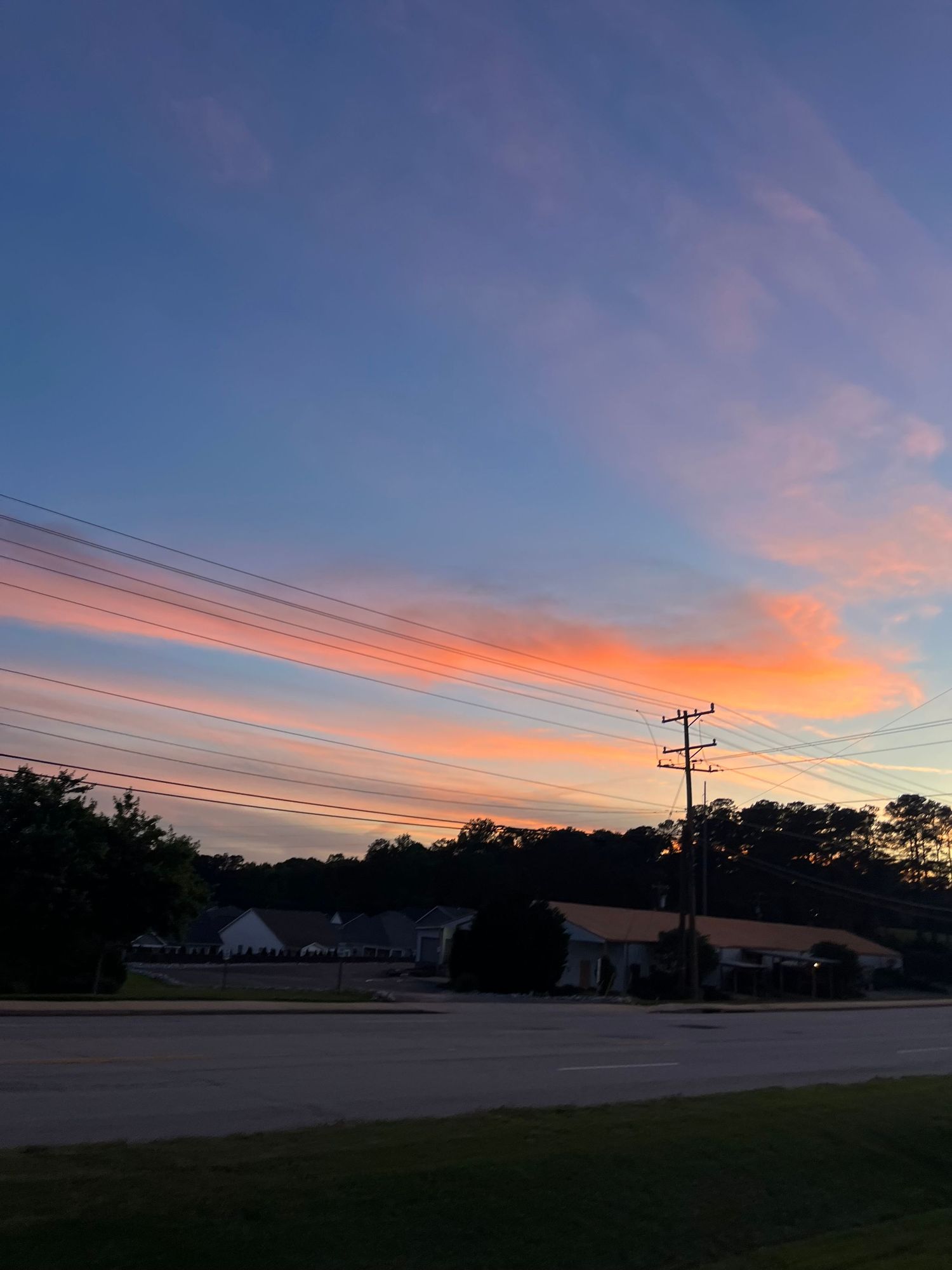 Vibrant orange and pink sunset sky stretches across a rural landscape with power lines.