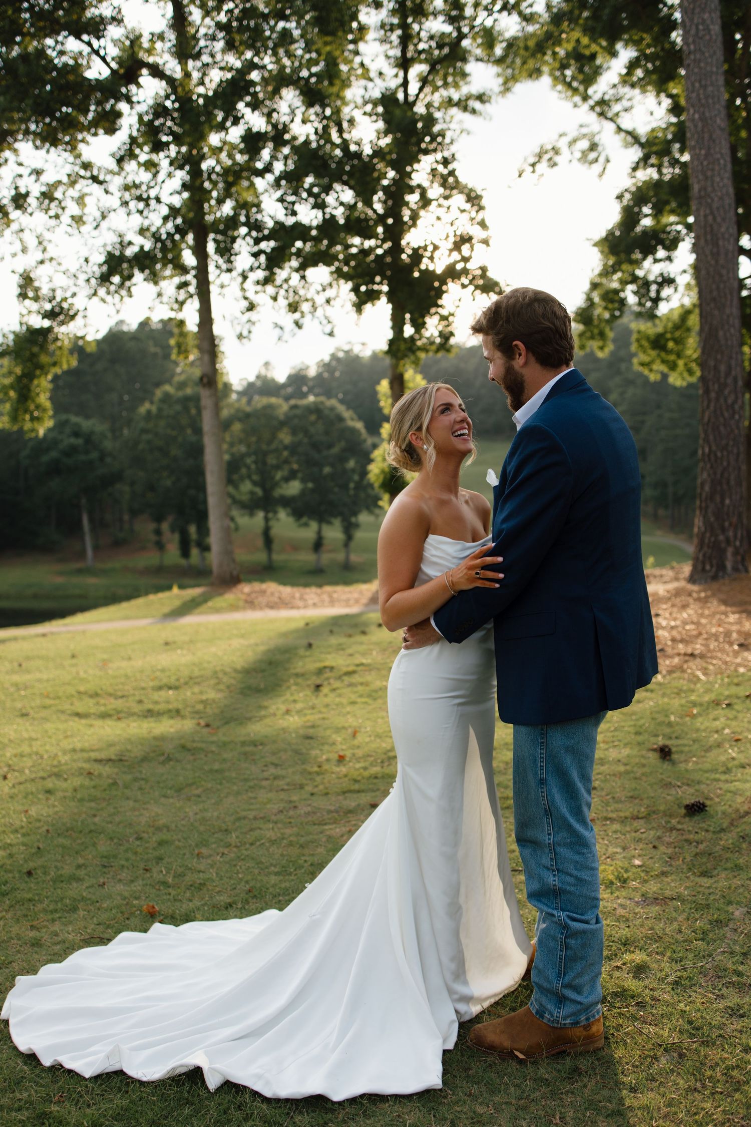 Couple shares intimate moment in elegant wedding attire against scenic wooded backdrop at sunset.