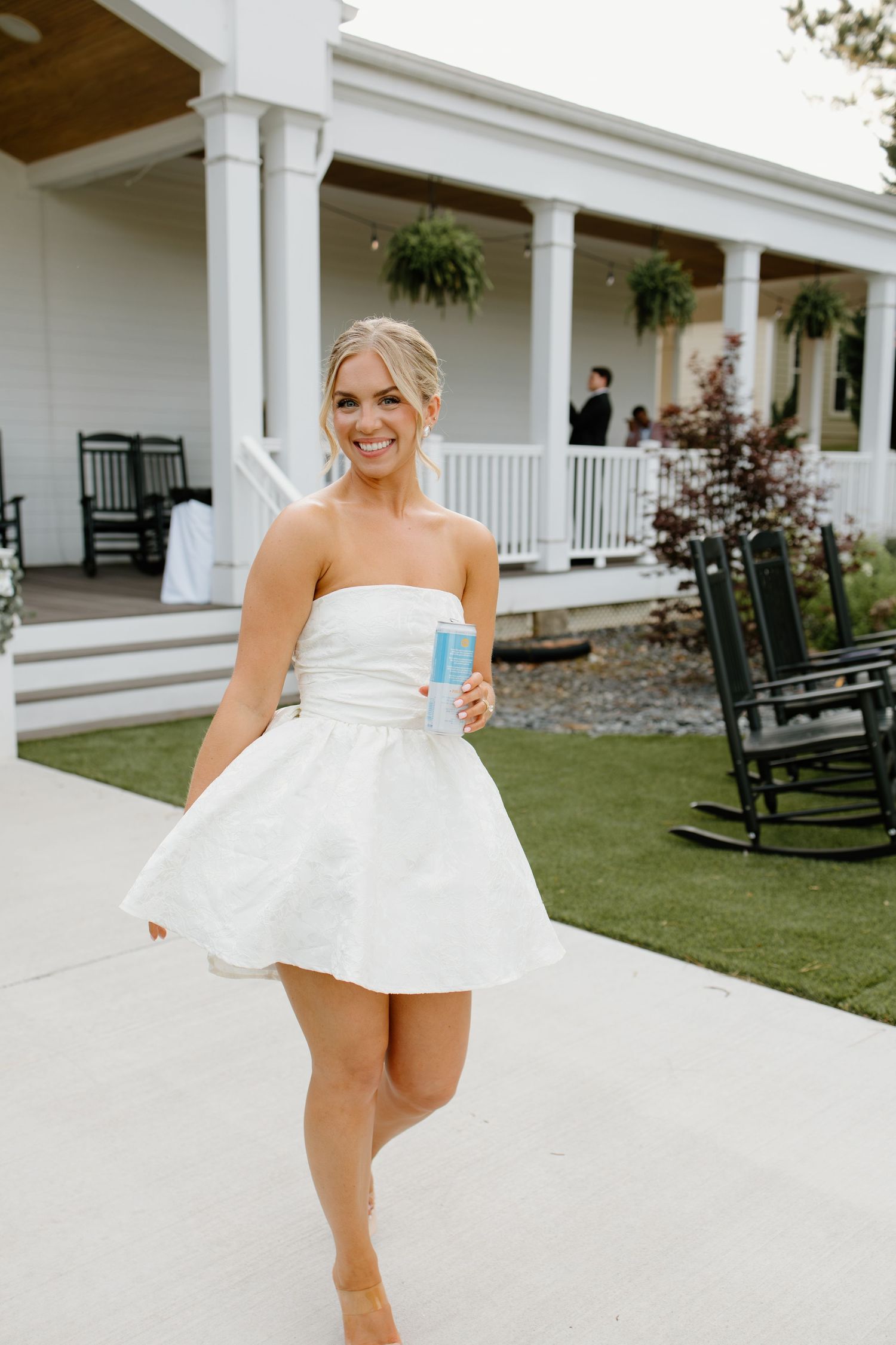 A woman in a white dress playfully twirls on a porch with white columns and rocking chairs.