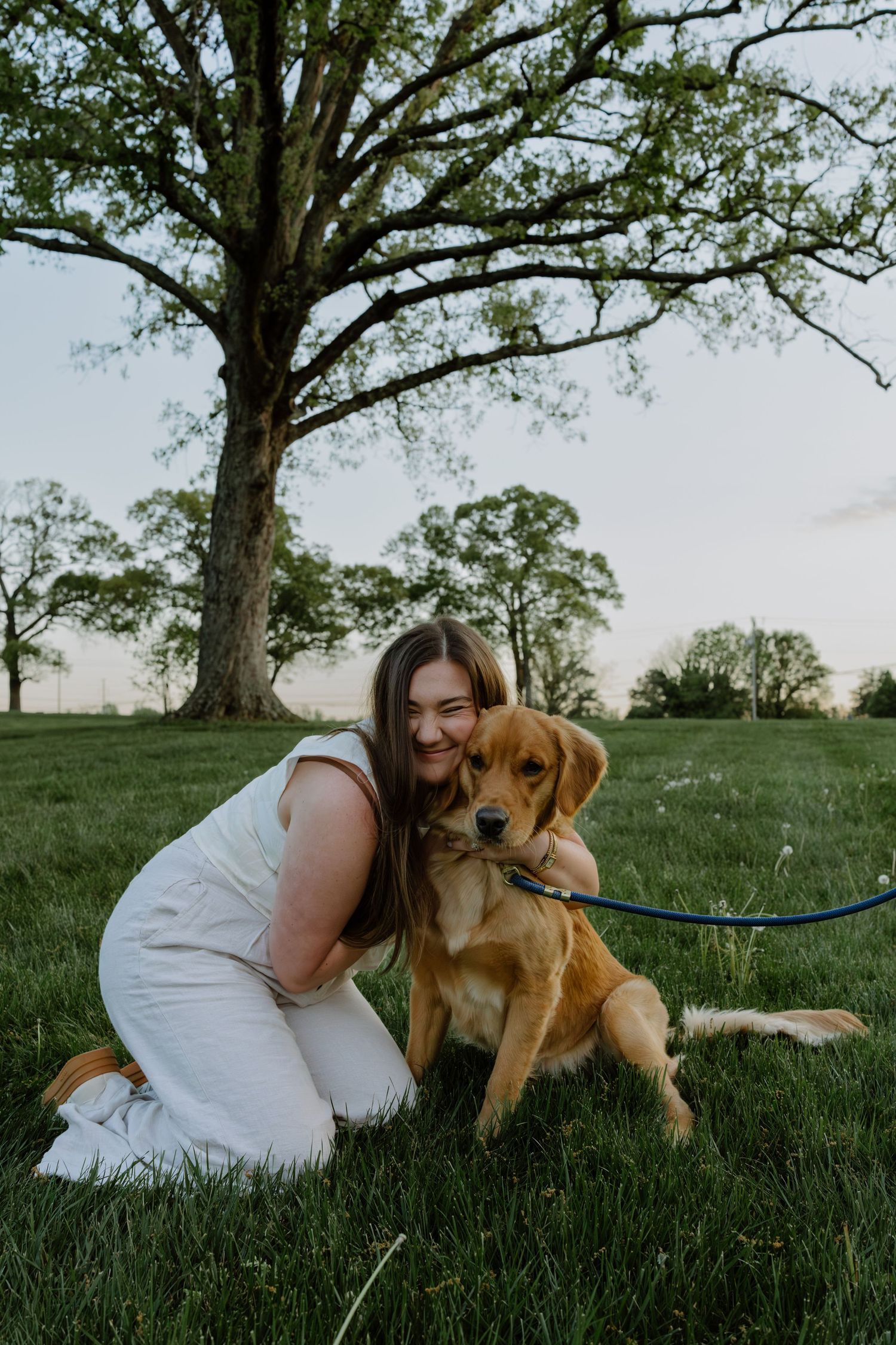 Young woman in white outfit hugging a golden retriever on green grass with a large tree at sunset.