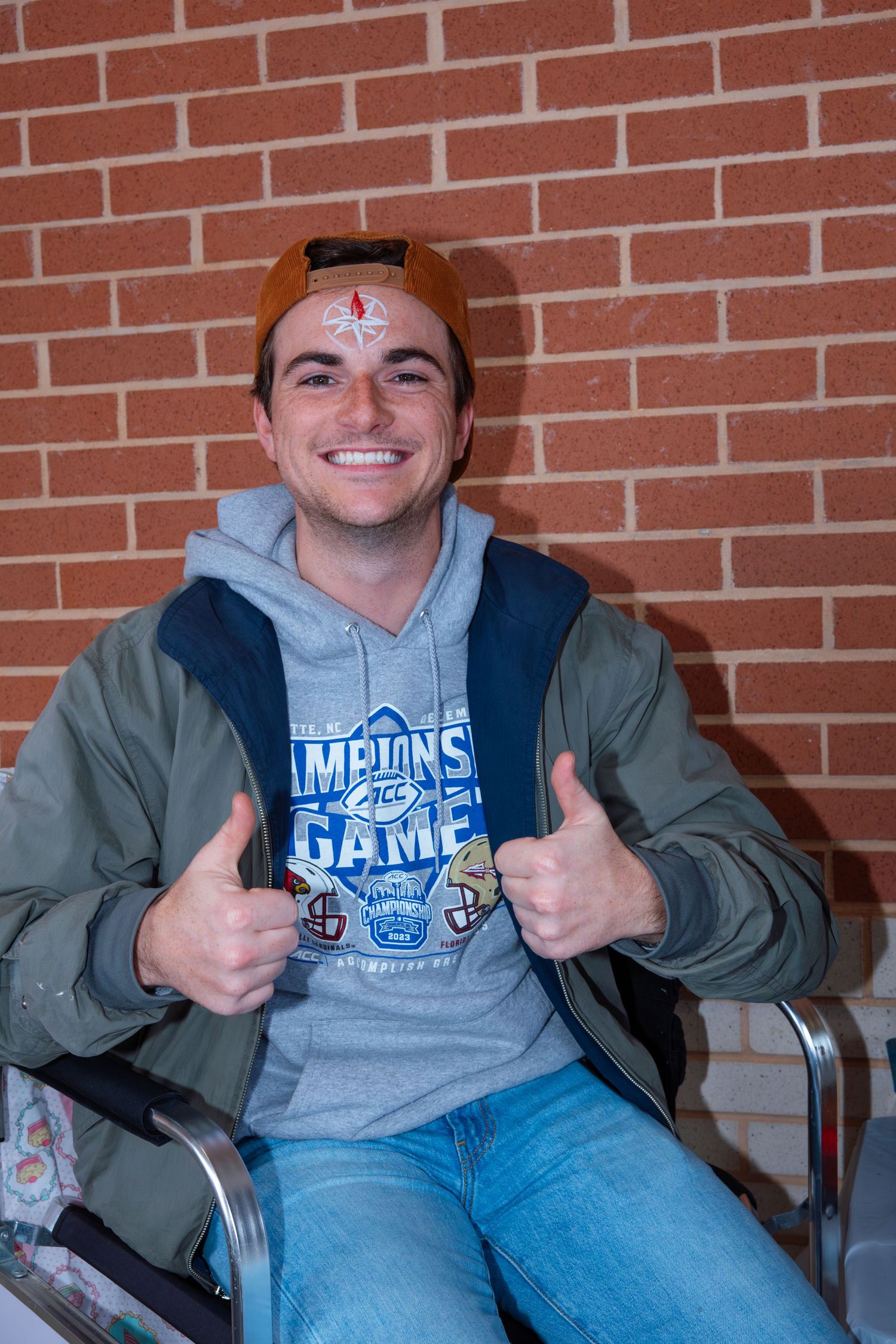 Student wearing a grey university sweatshirt gives thumbs up while sitting against brick wall.