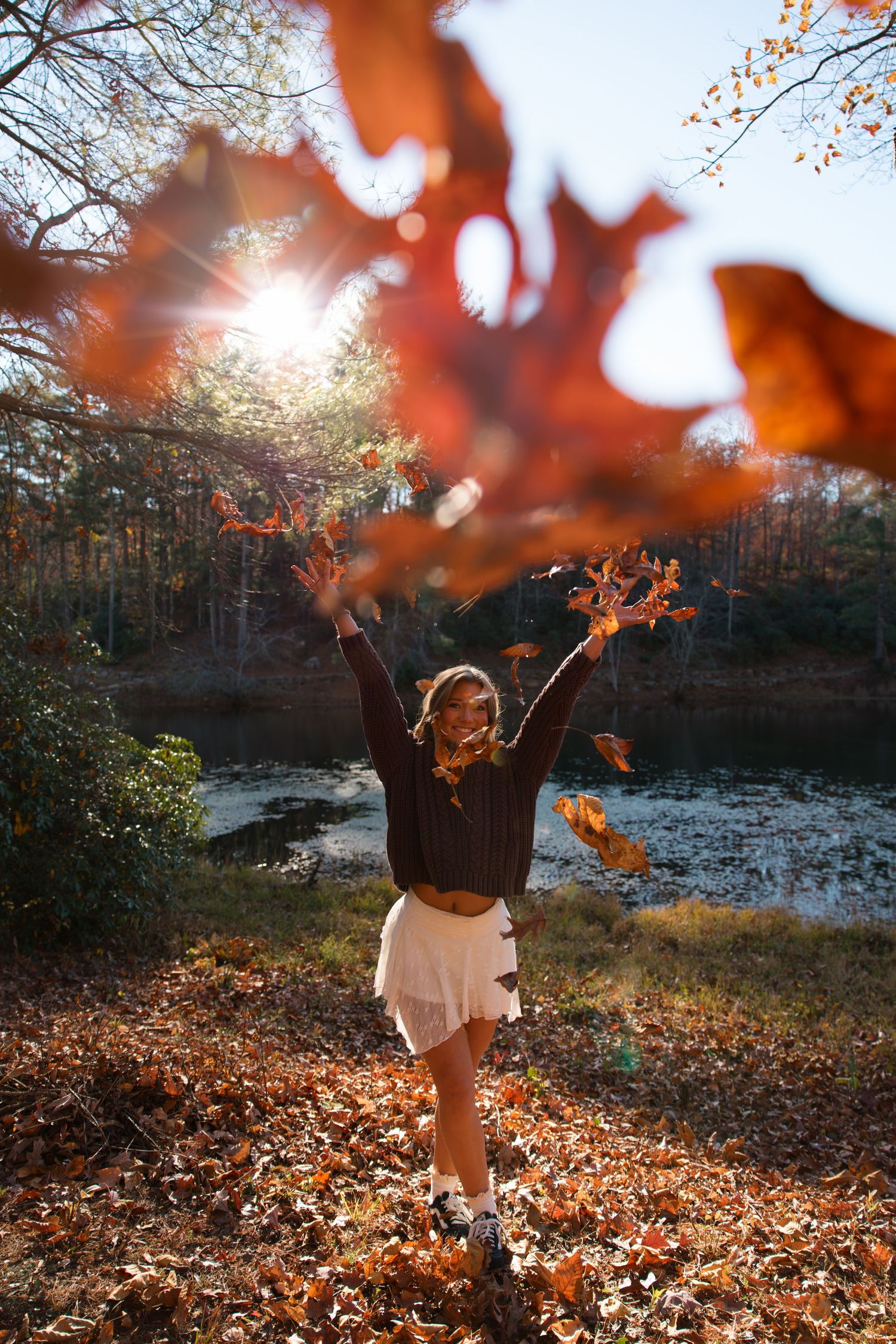 Person in a brown sweater twirls joyfully among autumn leaves with sunlight streaming through trees.