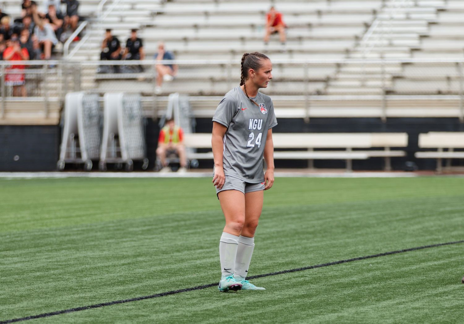 Soccer players lined up on the sideline during a match with stadium bleachers in background.