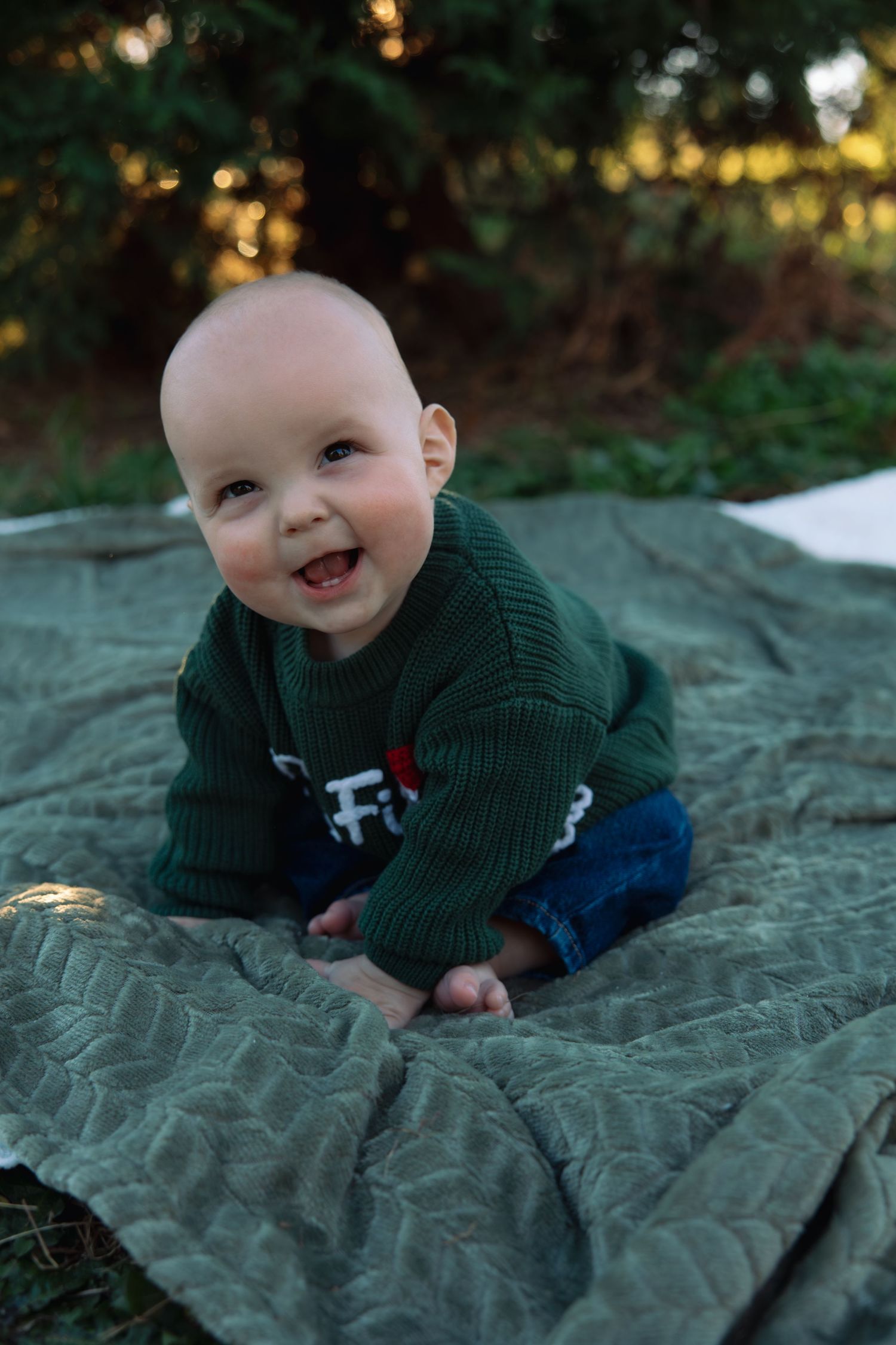 Baby in green sweater lying on blanket outdoors with joyful expression.