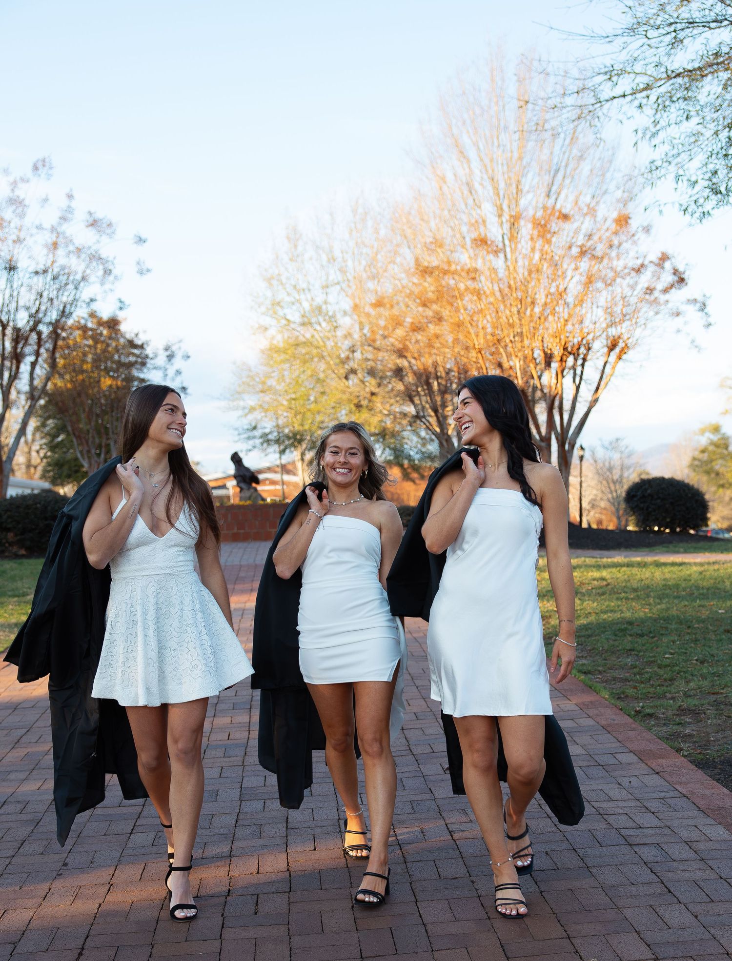Group of graduates in white dresses and black gowns walking together on brick pathway.