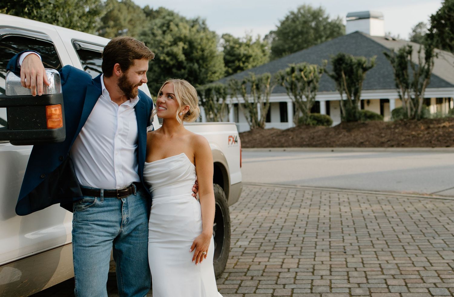 A couple in wedding attire leans against a white vehicle in a brick paved parking area.