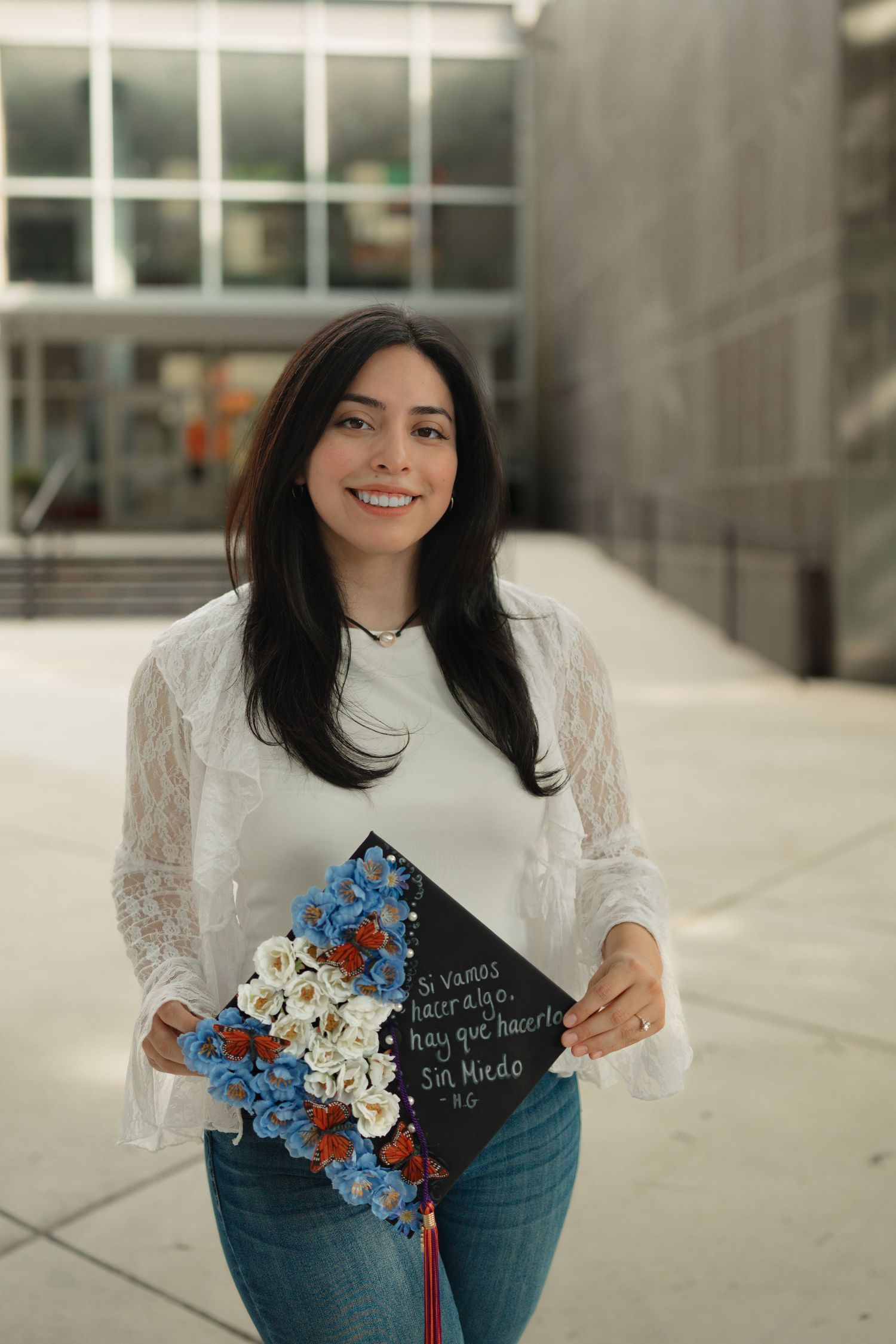 Smiling young woman holding a decorated black graduation cap with flowers and Spanish text, wearing white blouse and jeans.