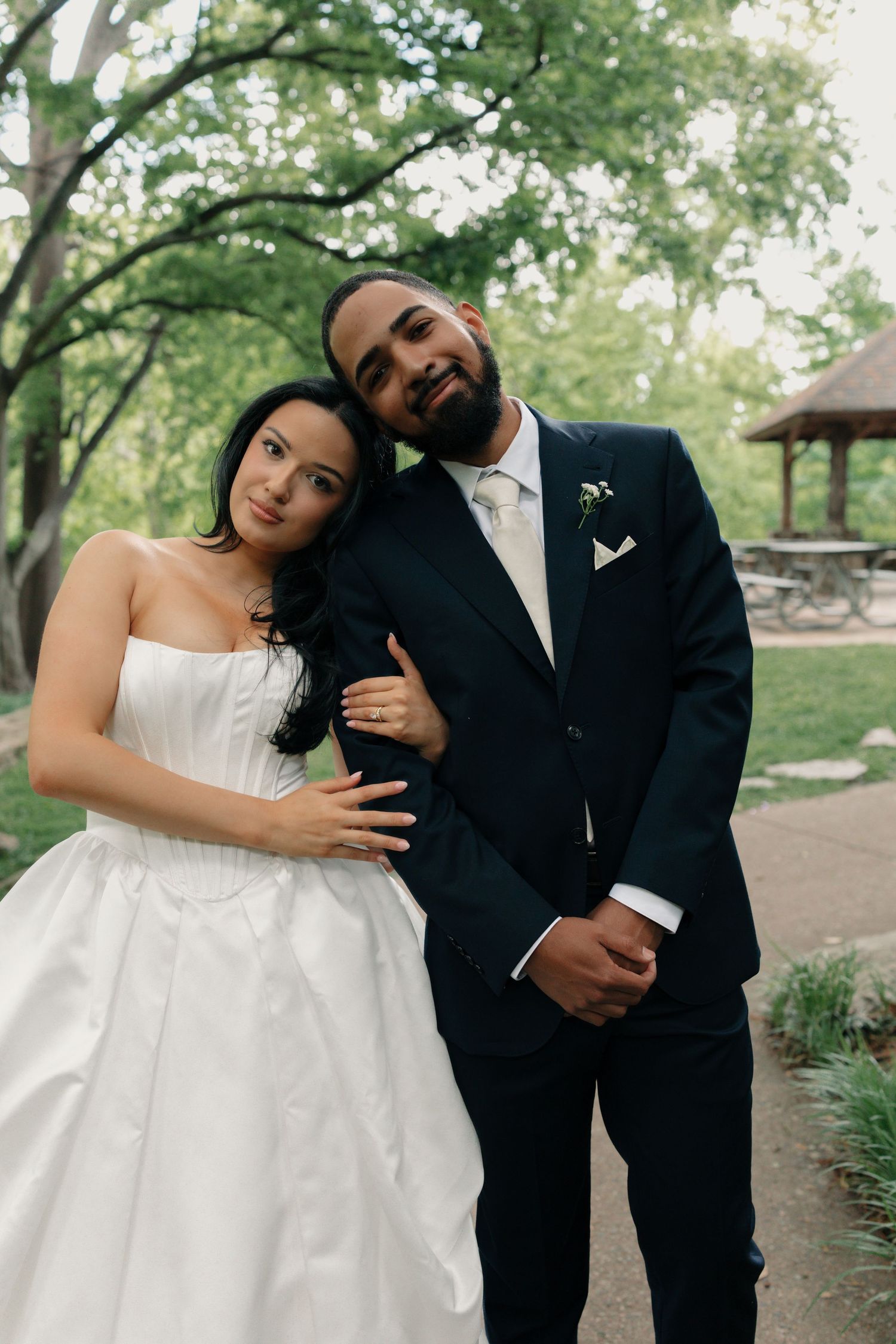 Bride in strapless white gown leans on groom in navy suit with boutonniere, posing in lush green park setting.