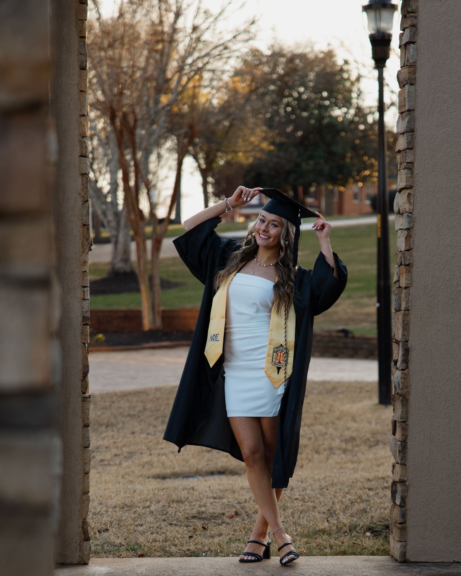 Graduate in cap and gown jumps playfully between pillars during outdoor photo session.