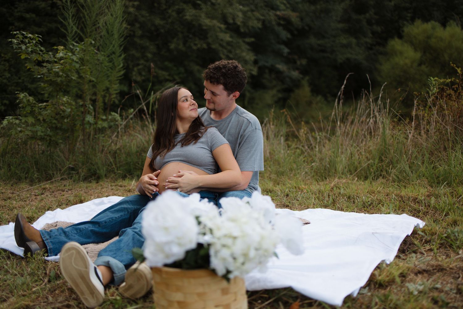 A couple in casual attire sits together on a blanket in a grassy outdoor setting during a romantic photoshoot.
