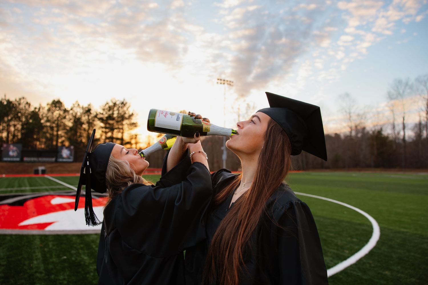 Graduates celebrating on a sports field at sunset while drinking from bottles.