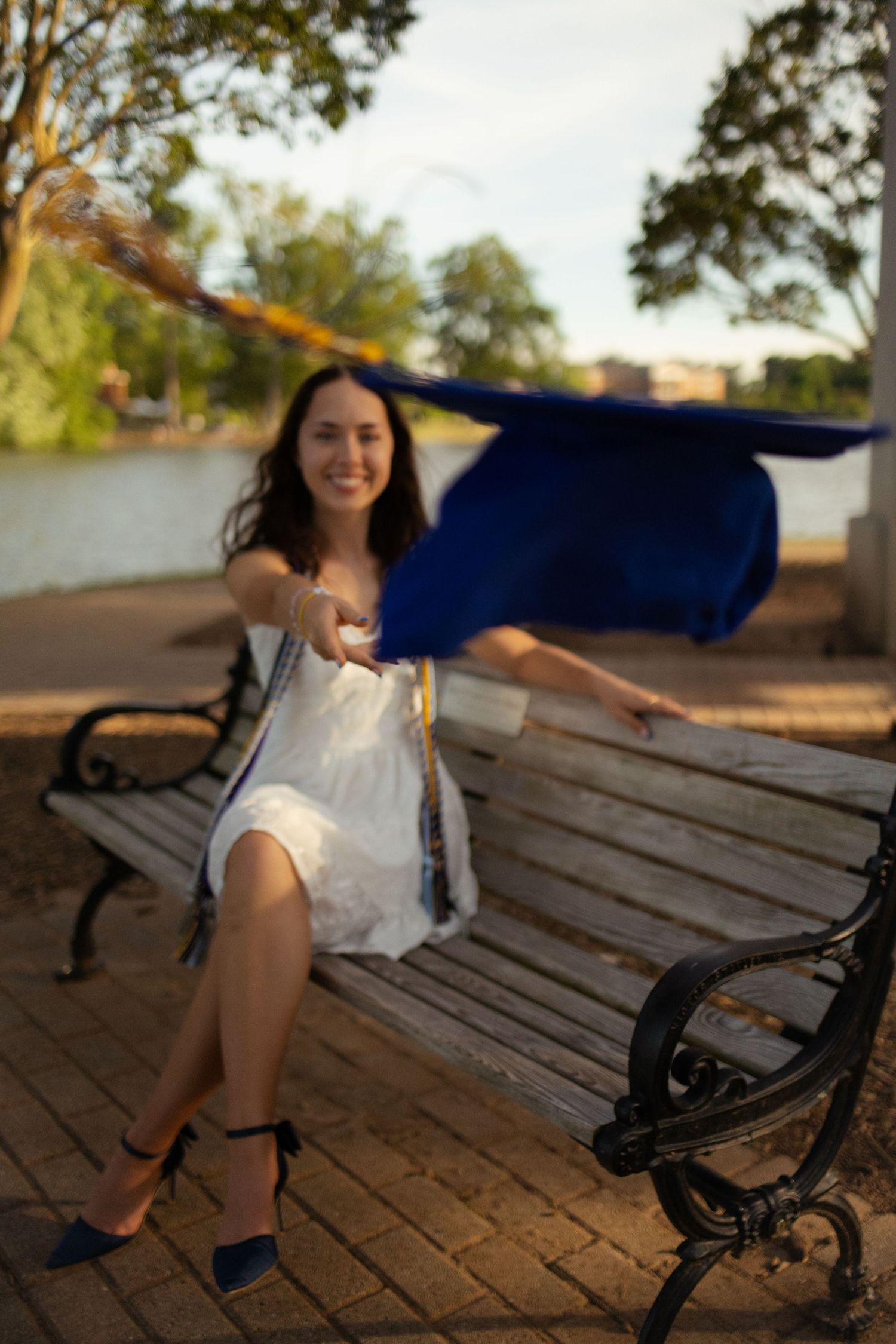 Graduate in white dress sitting on wooden bench during golden hour photography session.