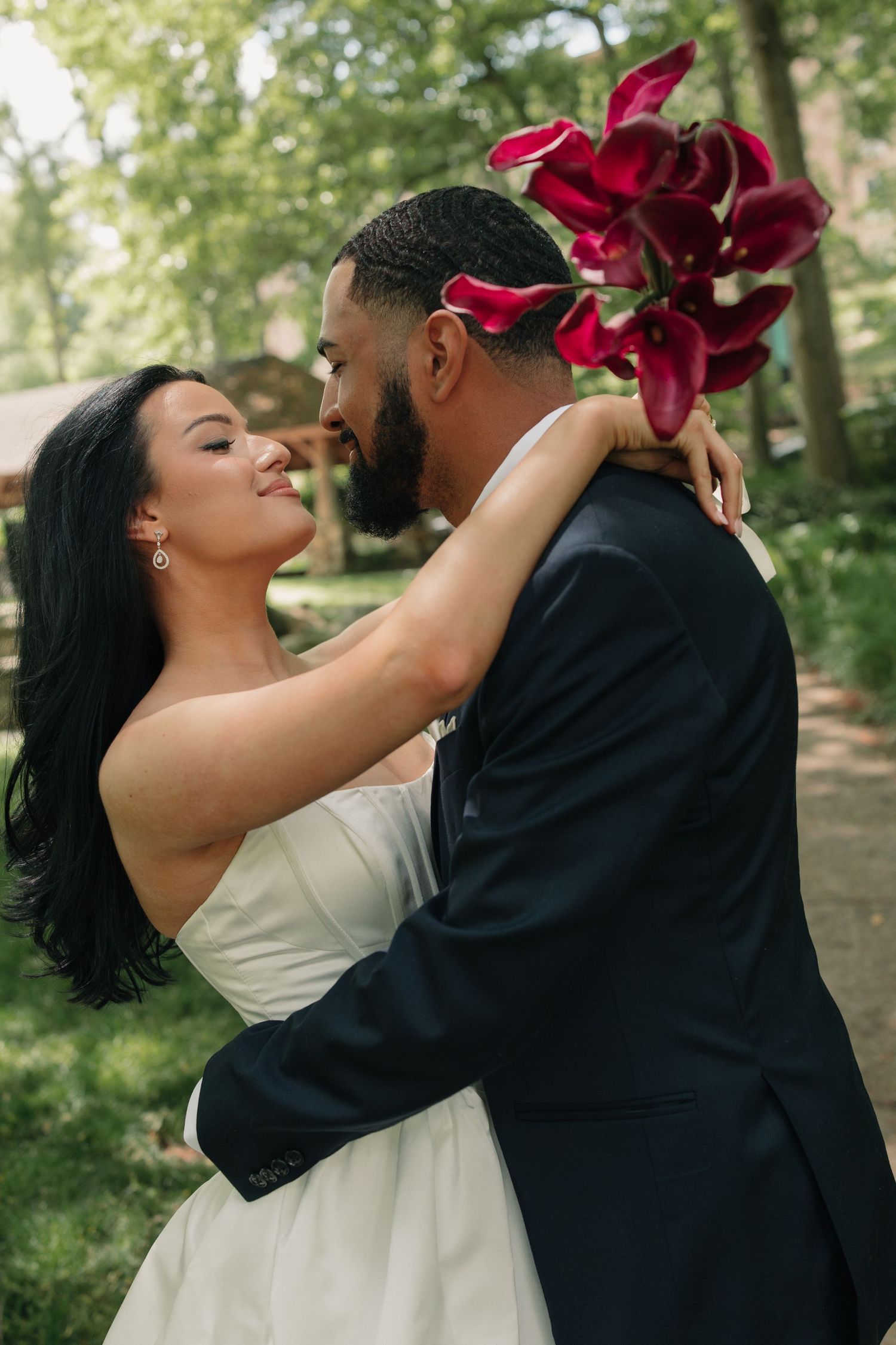 A bride and groom share a romantic embrace outdoors, with the bride holding red calla lilies while wearing a white strapless gown.