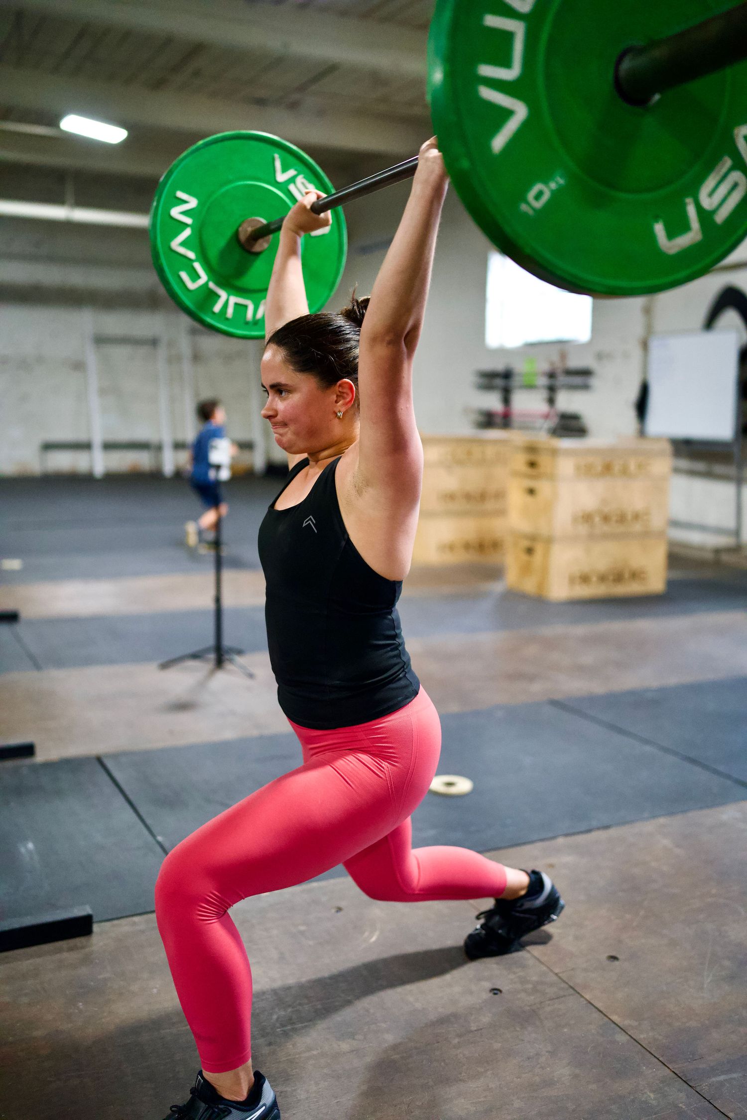 Woman performing an overhead squat lift with a Vulcan barbell in a CrossFit gym wearing pink leggings.