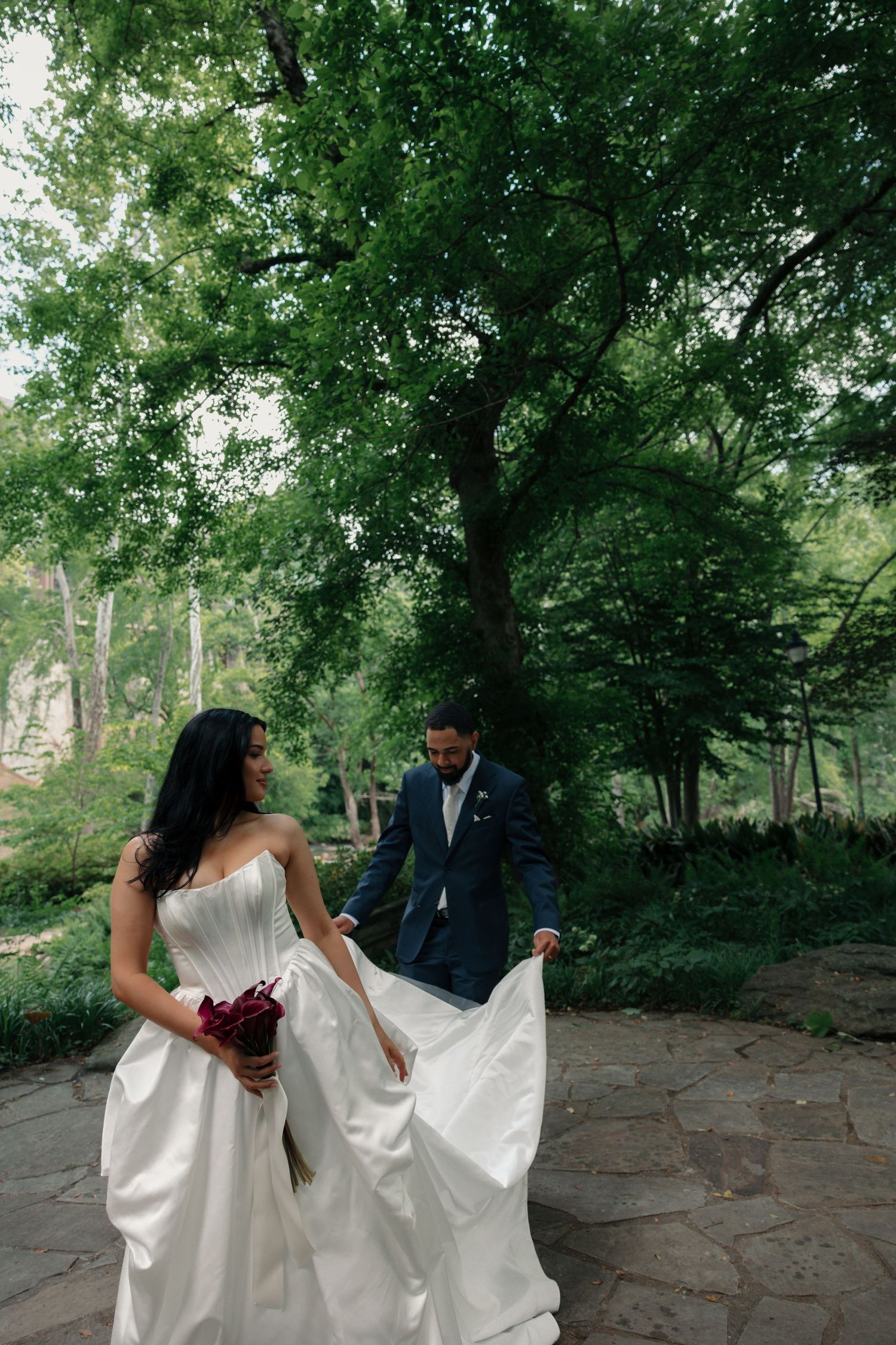 Bride in white strapless gown holding bouquet with groom in navy suit walking on stone path in lush green park.