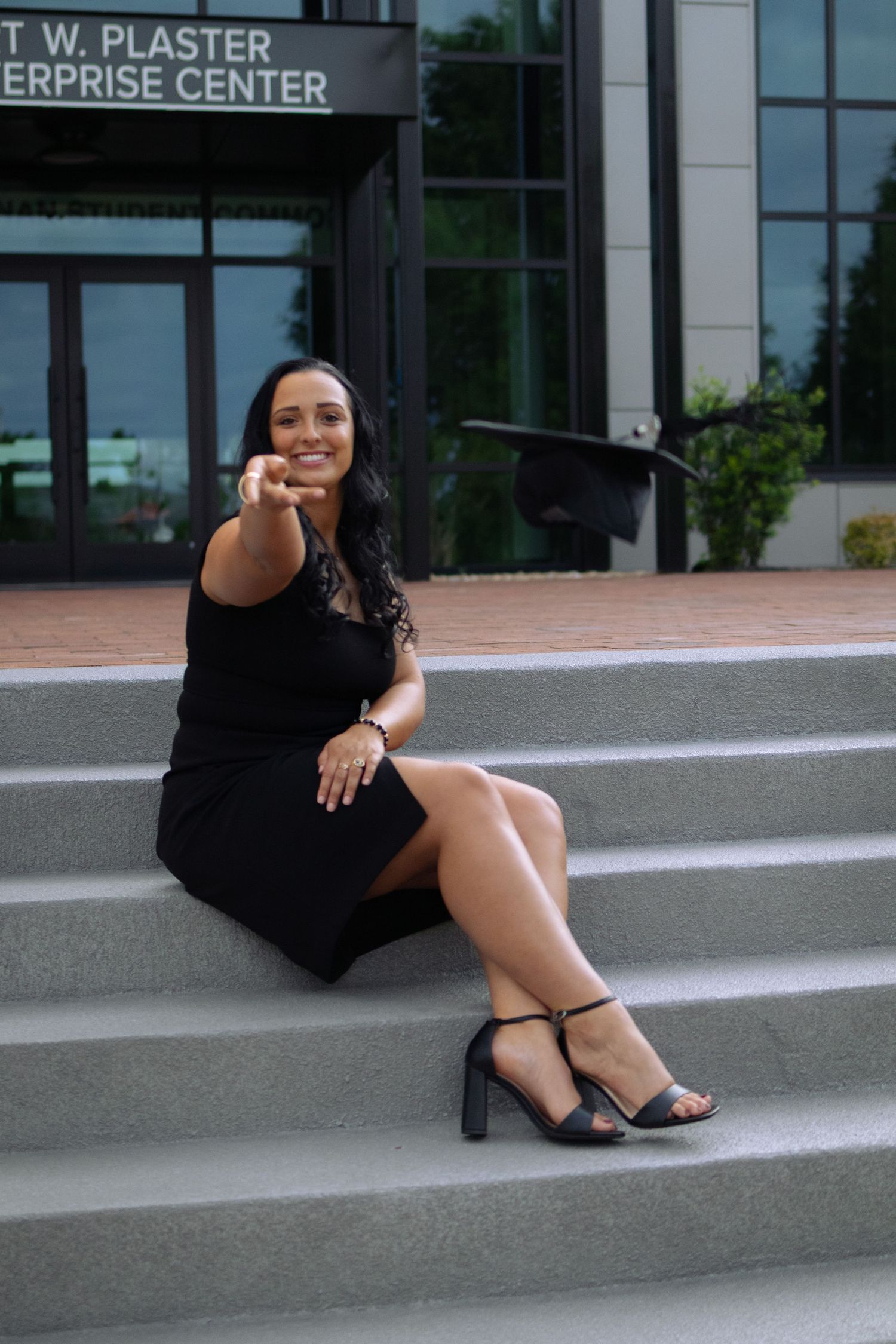 Graduate sitting on concrete steps outside T W Plaster building in black dress and heels.