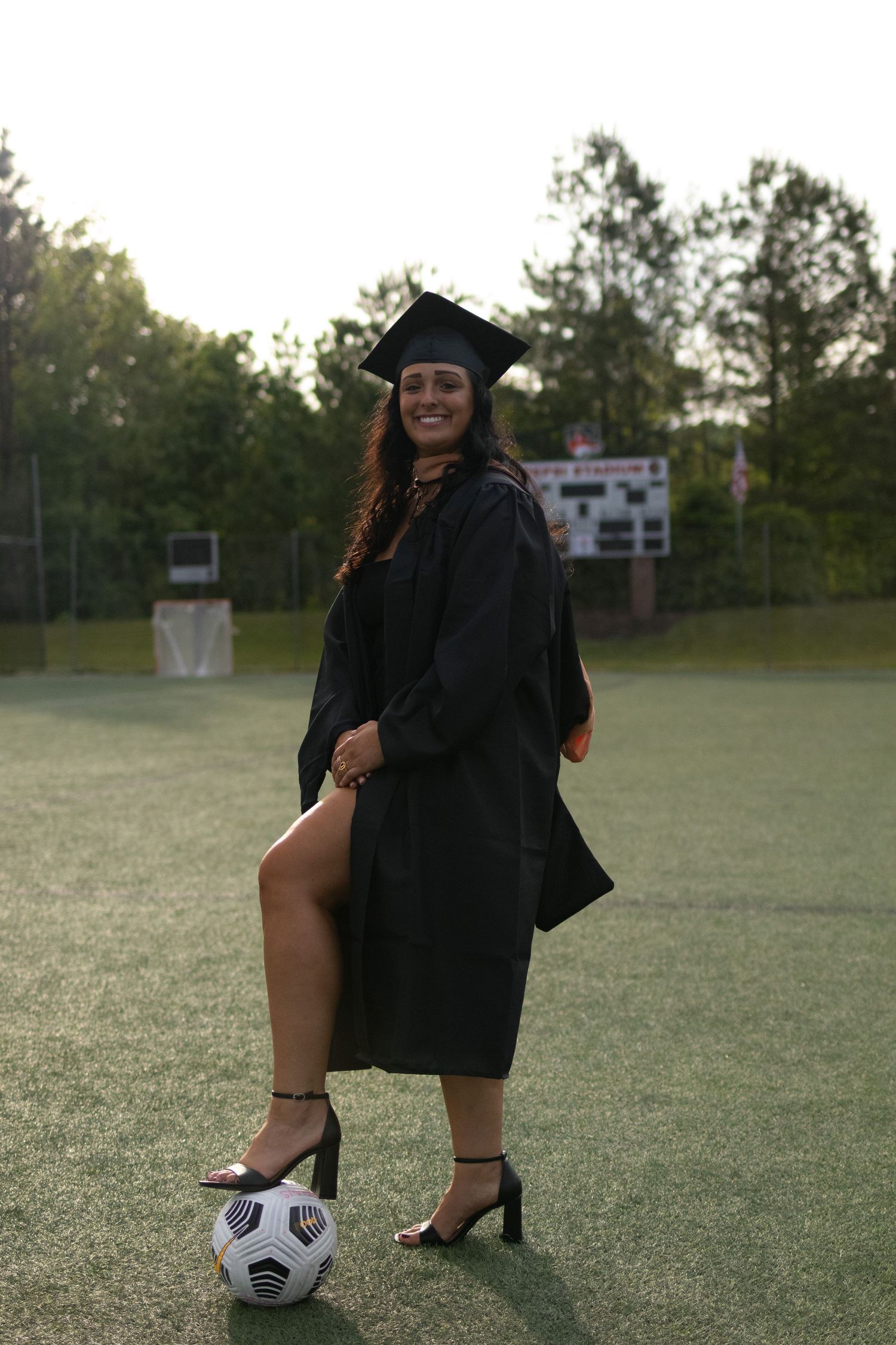 Graduate in cap and gown posing with soccer ball on sports field during sunset.