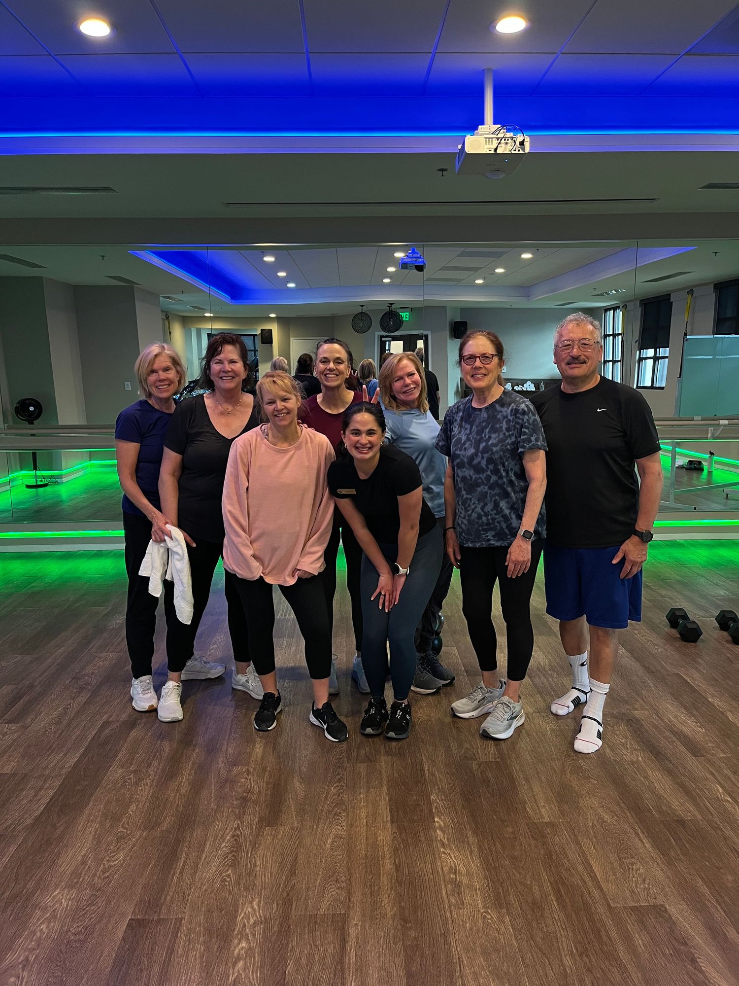 Group of adults posing together in a fitness studio with green and blue LED lighting after a workout class.