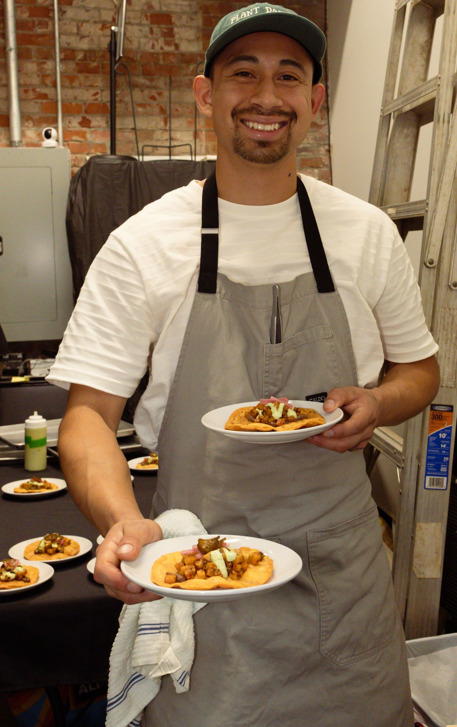 Chef wearing gray apron presents beautifully plated dishes in rustic brick-walled kitchen.