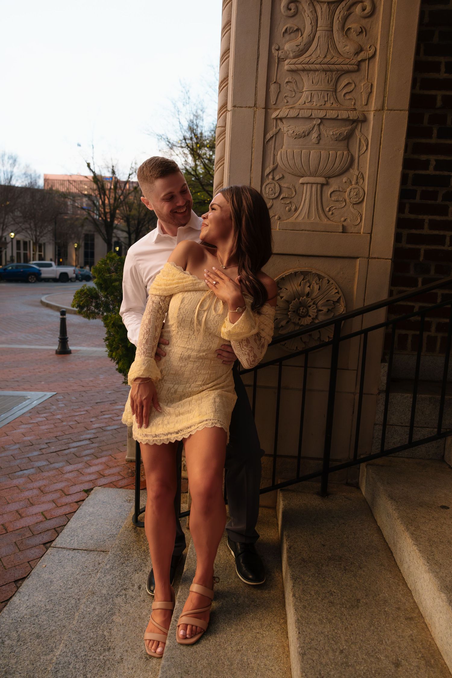A couple poses romantically by an ornate stone building column on a brick plaza at dusk.
