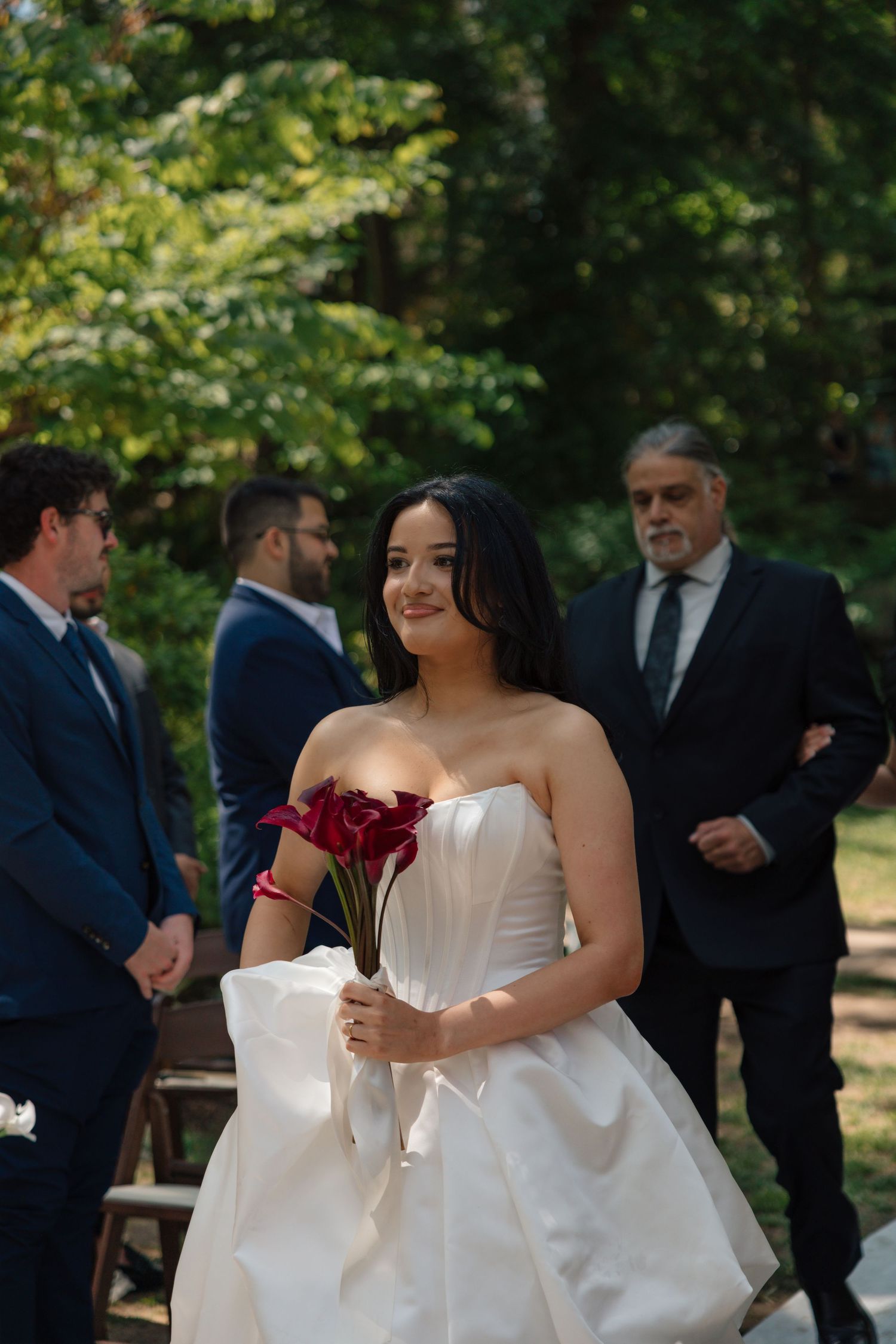Bride in strapless white gown holding red calla lilies walks down outdoor aisle with guests in suits behind her.
