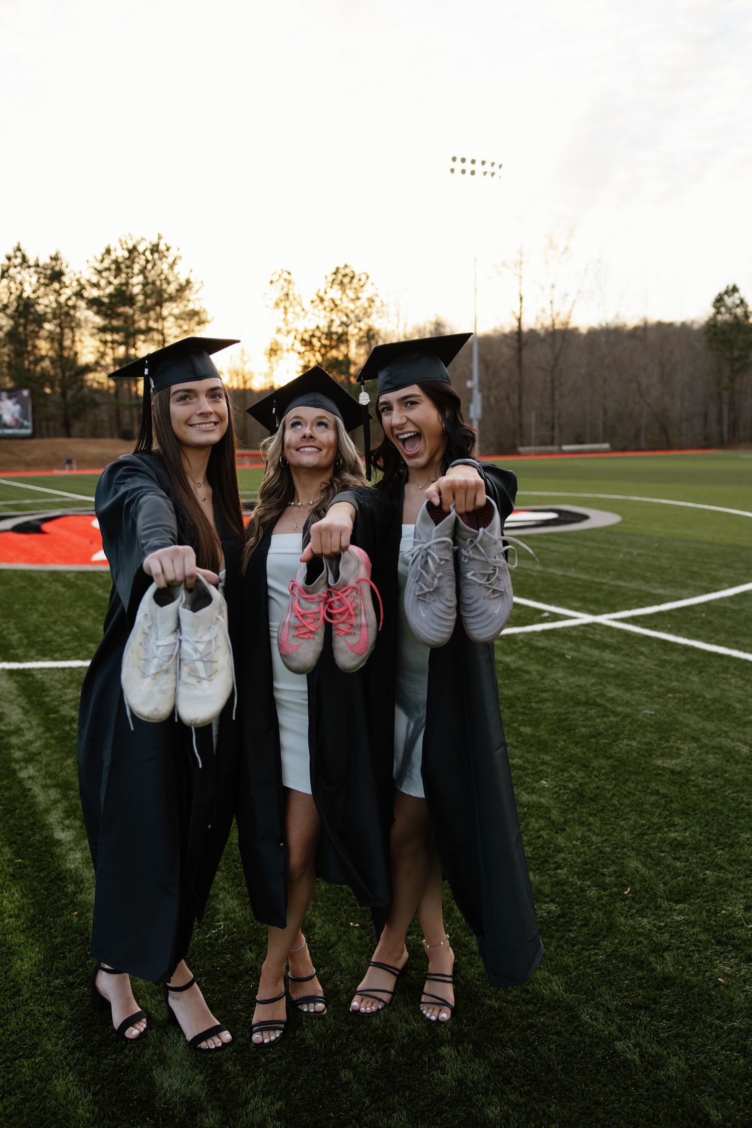 Graduates in black gowns pose with pink shoes on a football field during a celebratory photoshoot.