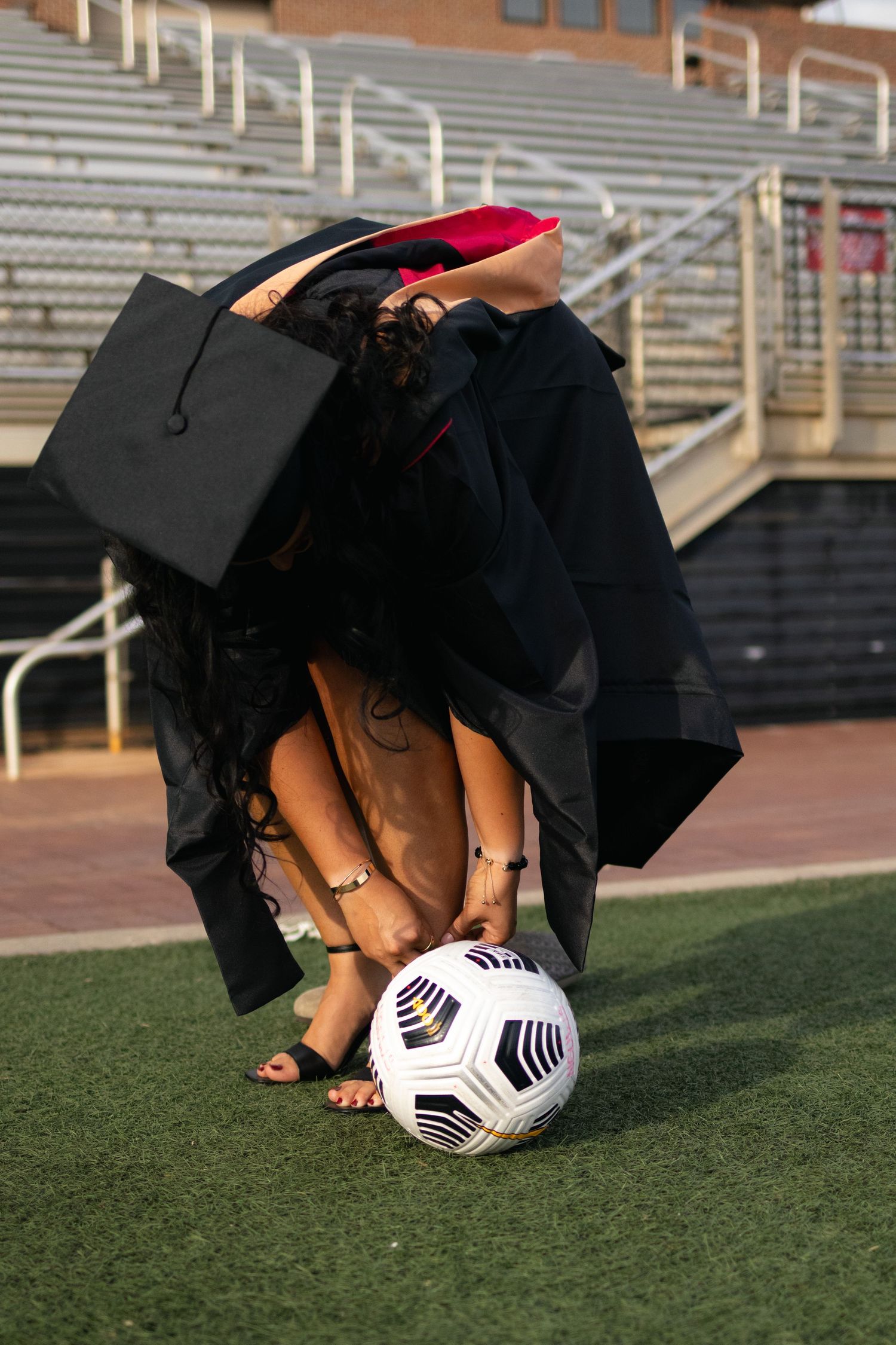 Soccer ball on artificial turf with person in black dress bending down near stadium bleachers.