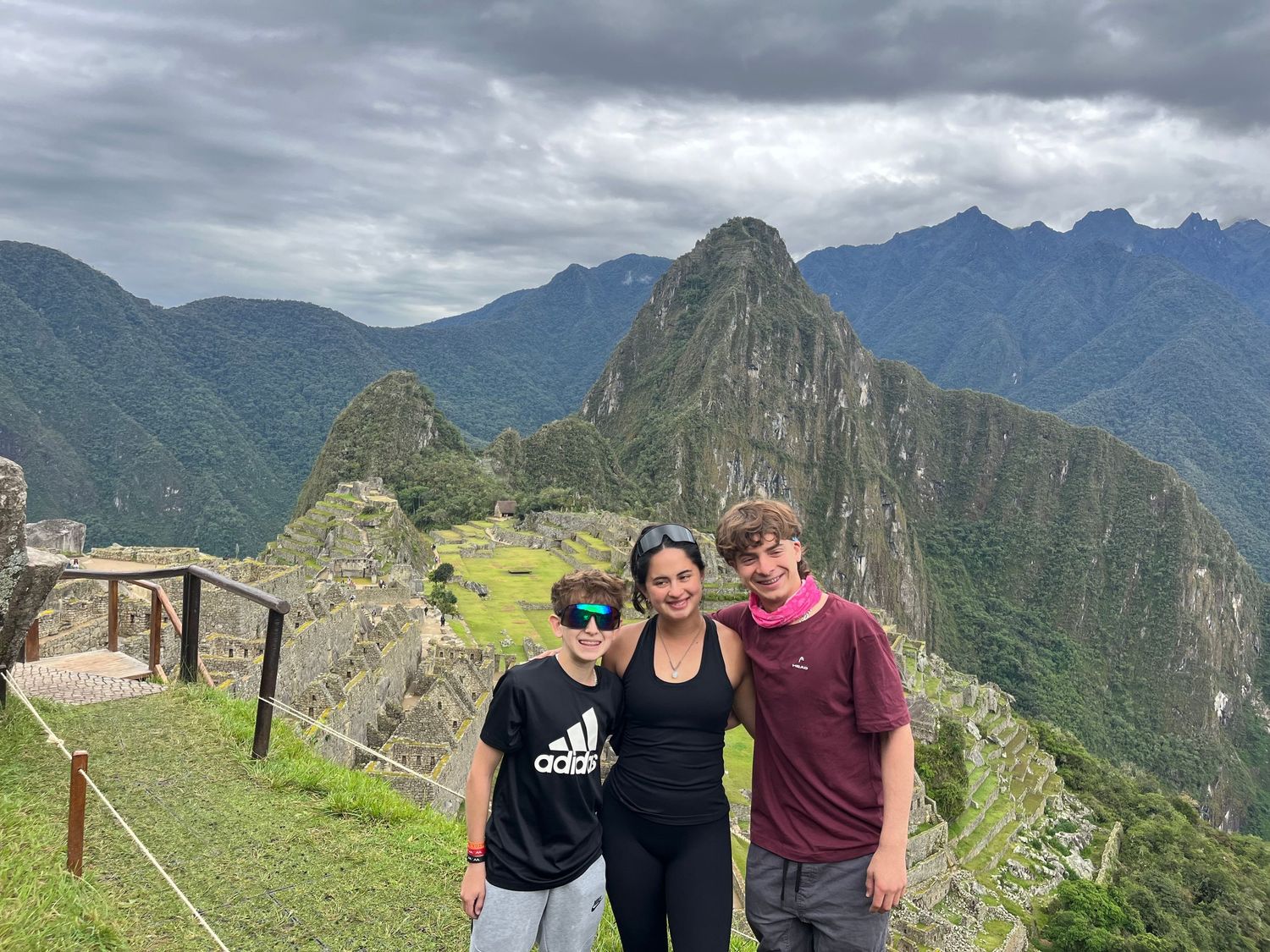 Three tourists pose smiling at Machu Picchu, Peru, with ancient Incan ruins and misty mountain peaks in the background.