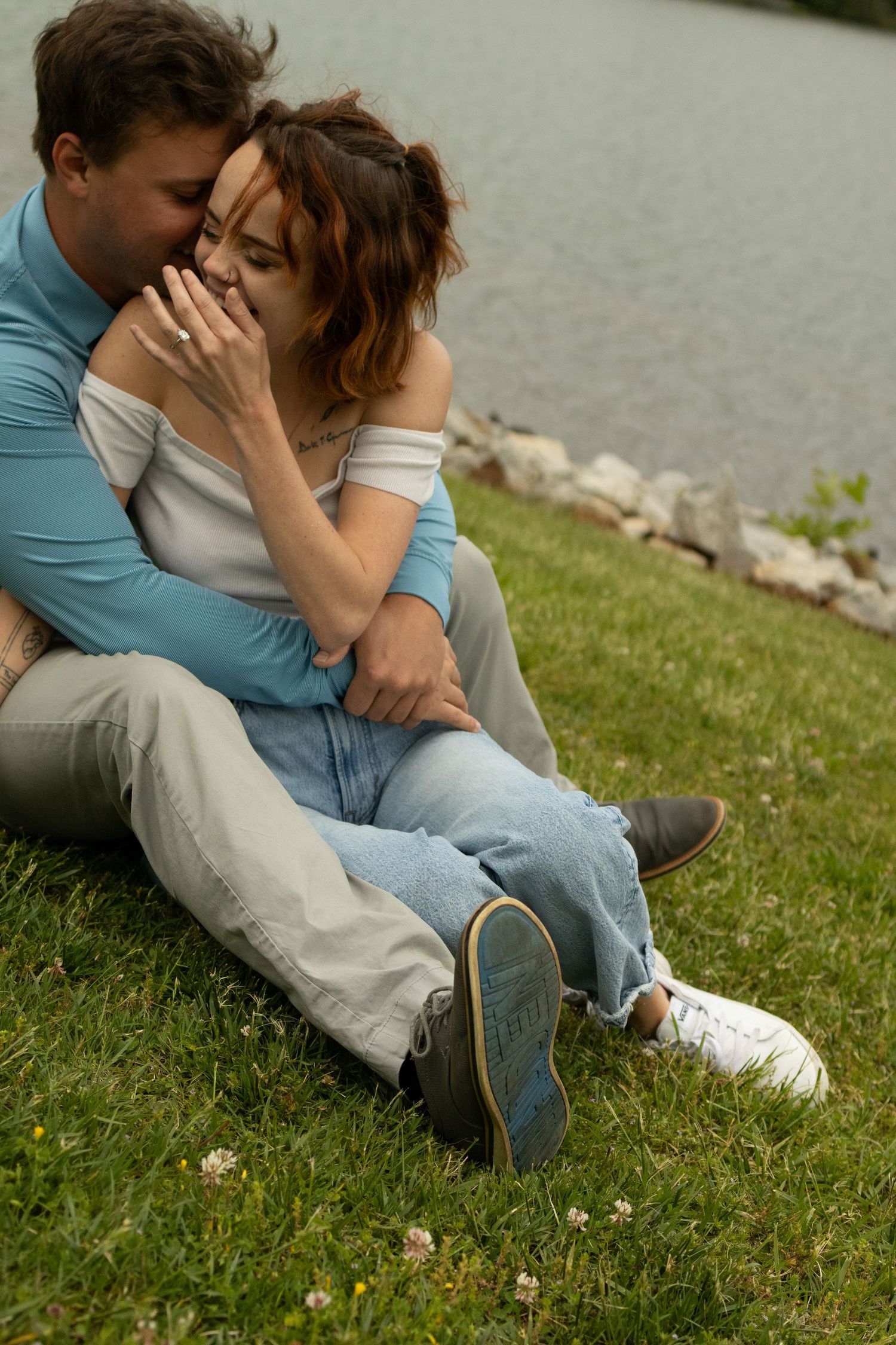A couple relaxes on grassy lakeside ground wearing casual attire.