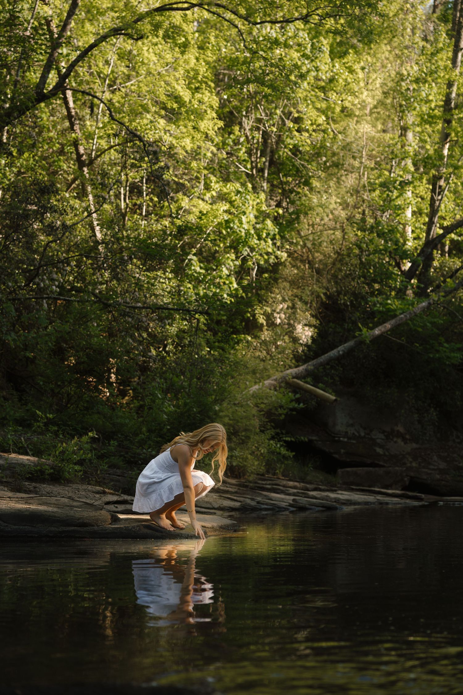 Woman in white dress crouching by a tranquil forest stream, her reflection visible in the calm water surrounded by lush green trees.