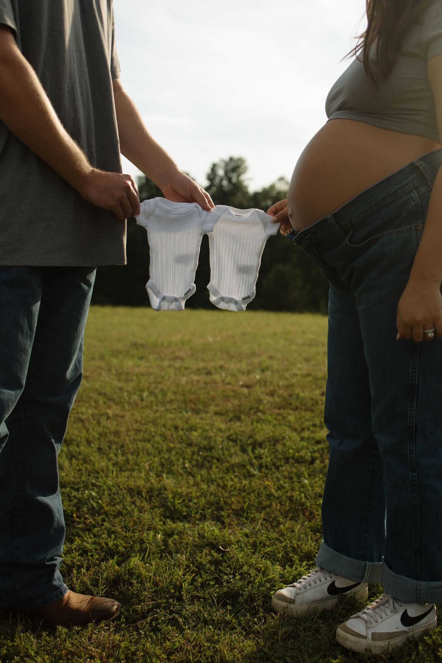 Couple holding tiny baby outfits in grassy field during pregnancy announcement.