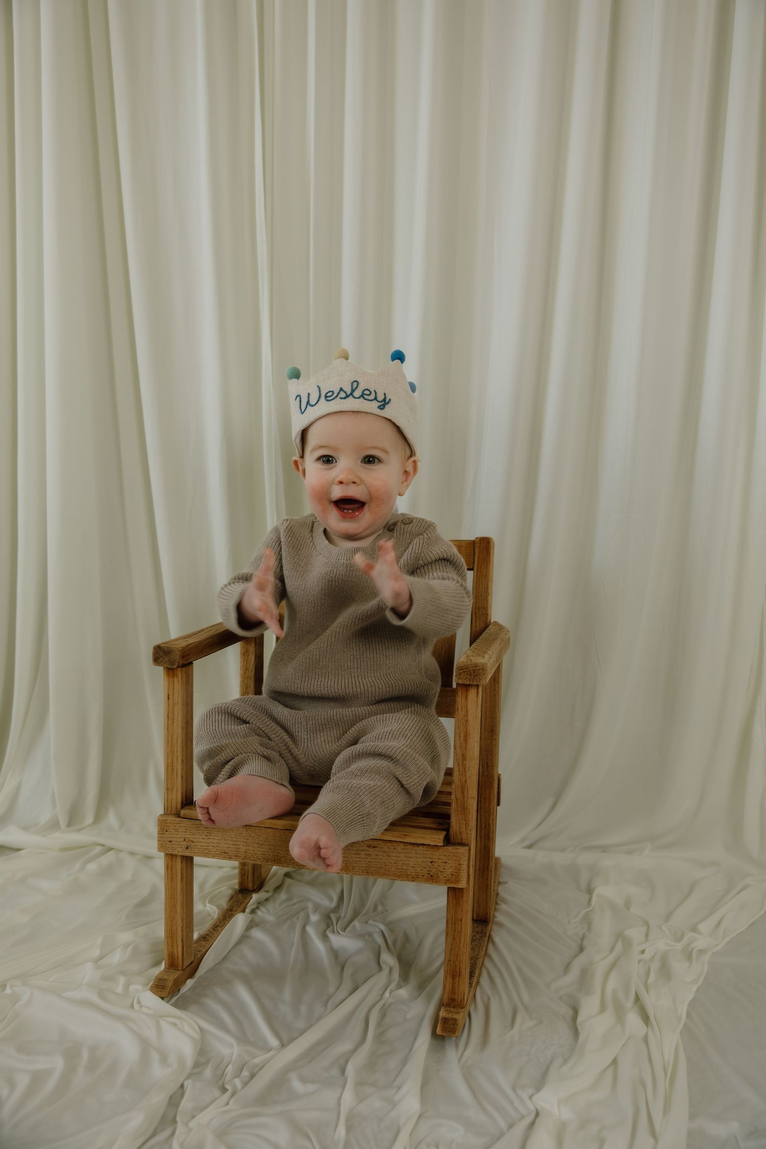 Smiling baby wearing crown sits in wooden rocking chair against white backdrop.