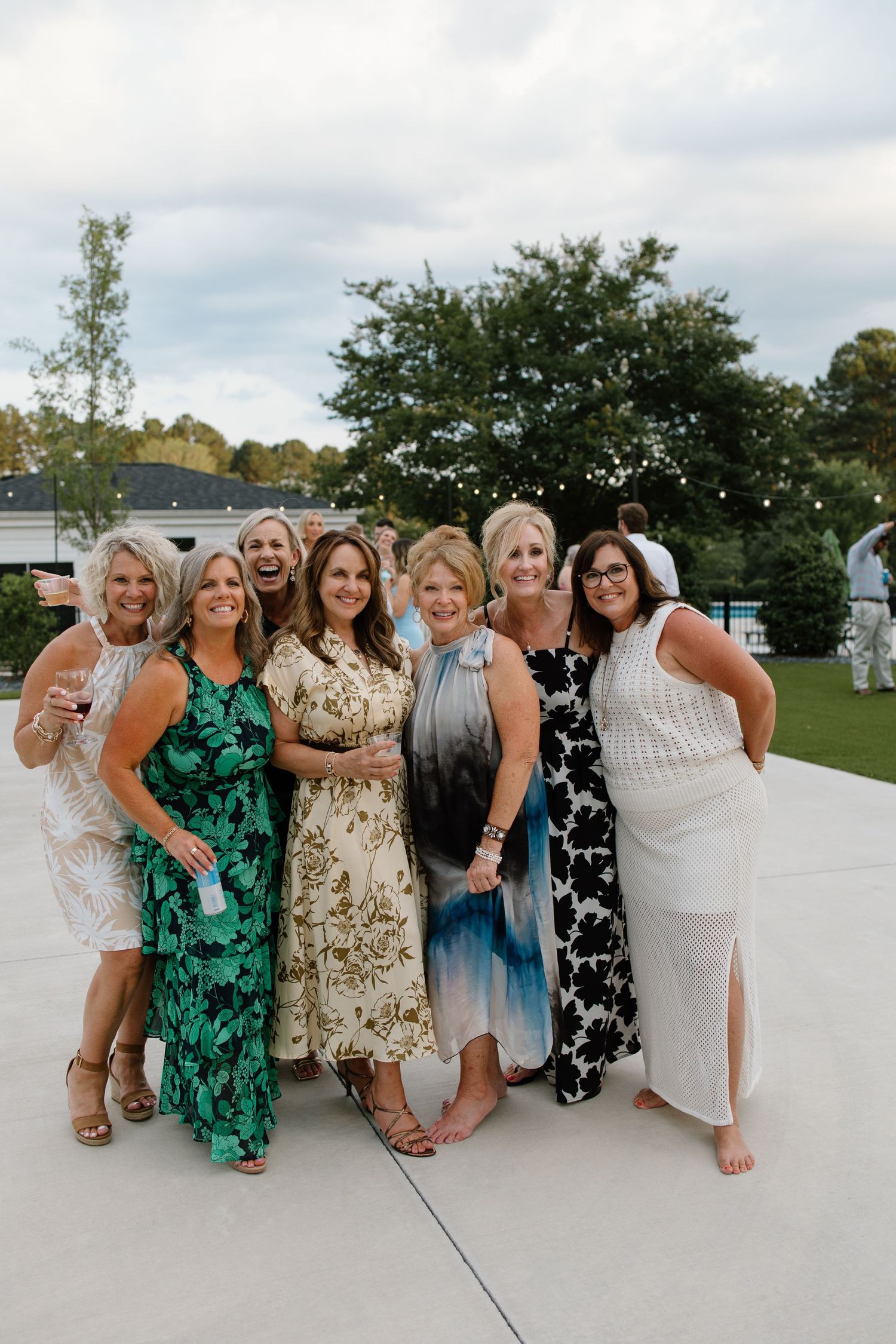A group of friends in colorful dresses pose together on a white concrete surface outdoors.