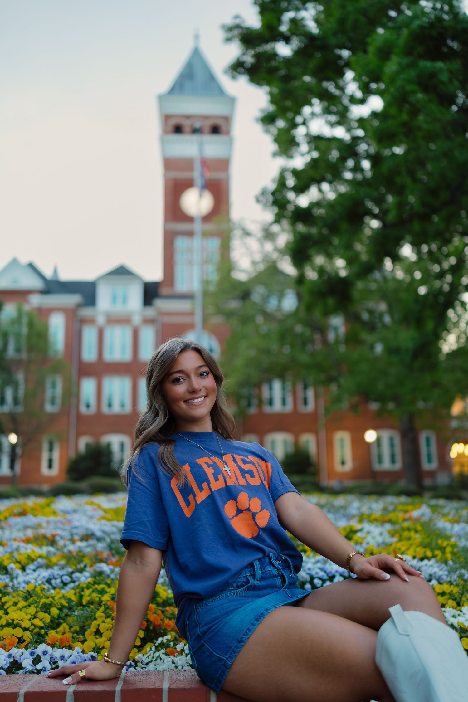 Young woman in Clemson University blue t-shirt sitting by colorful flowers with brick clock tower behind her.