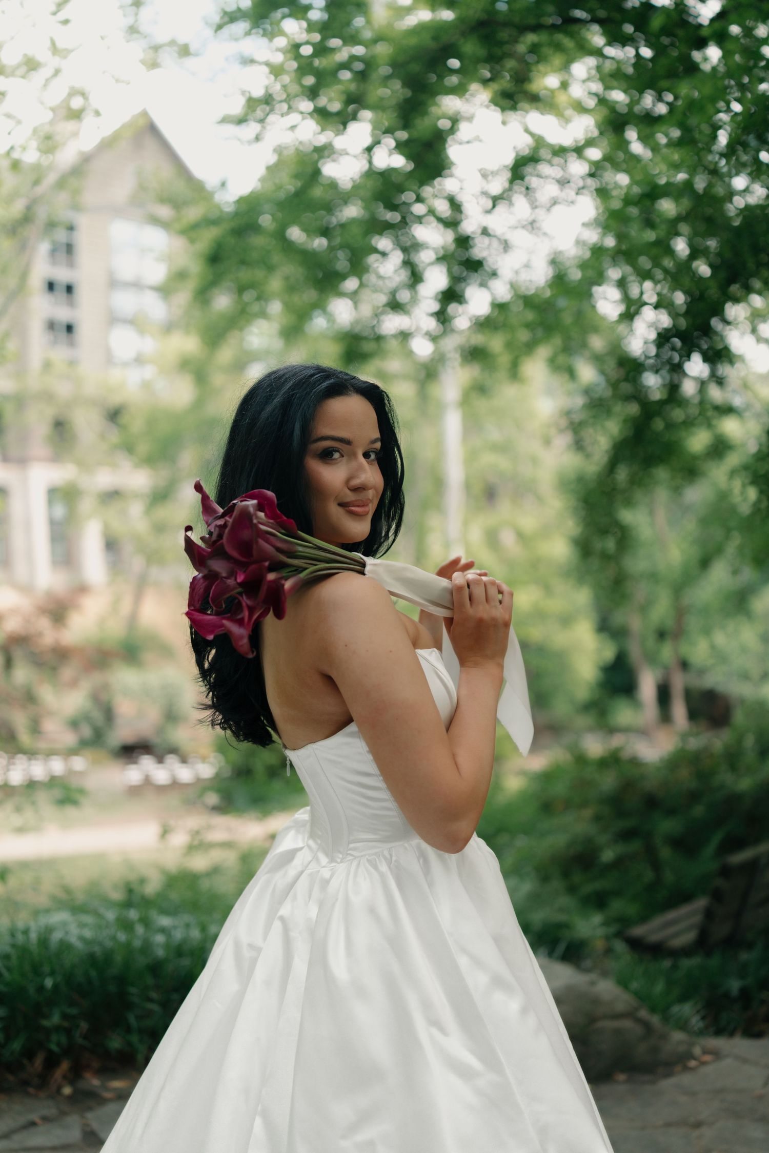 Bride in white strapless gown holds dark red calla lily bouquet over shoulder, smiling in lush green garden setting.