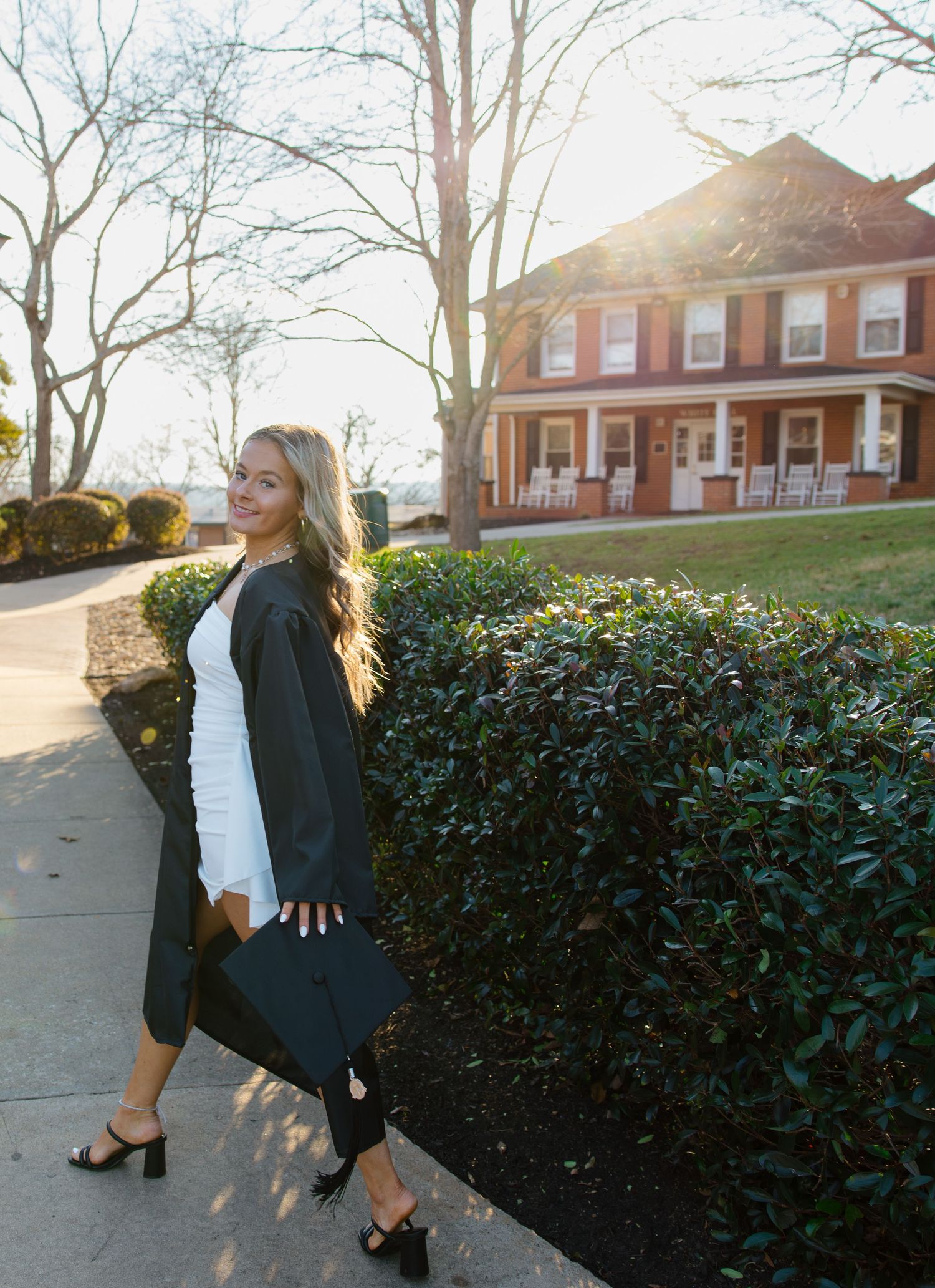 Graduate in black gown poses confidently on campus pathway during sunset.