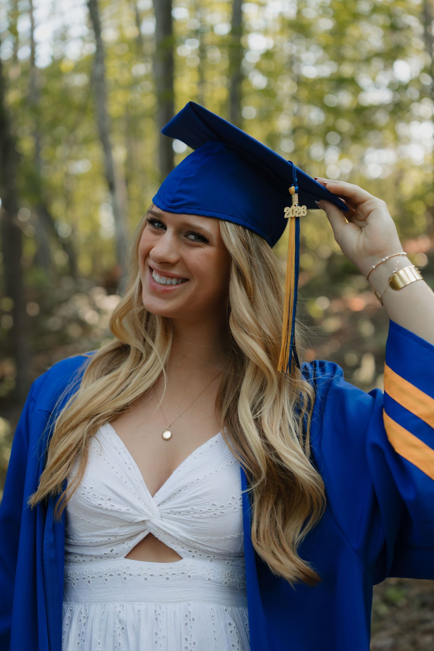 Smiling blonde woman in blue graduation cap and gown with gold tassel posing outdoors among autumn trees.