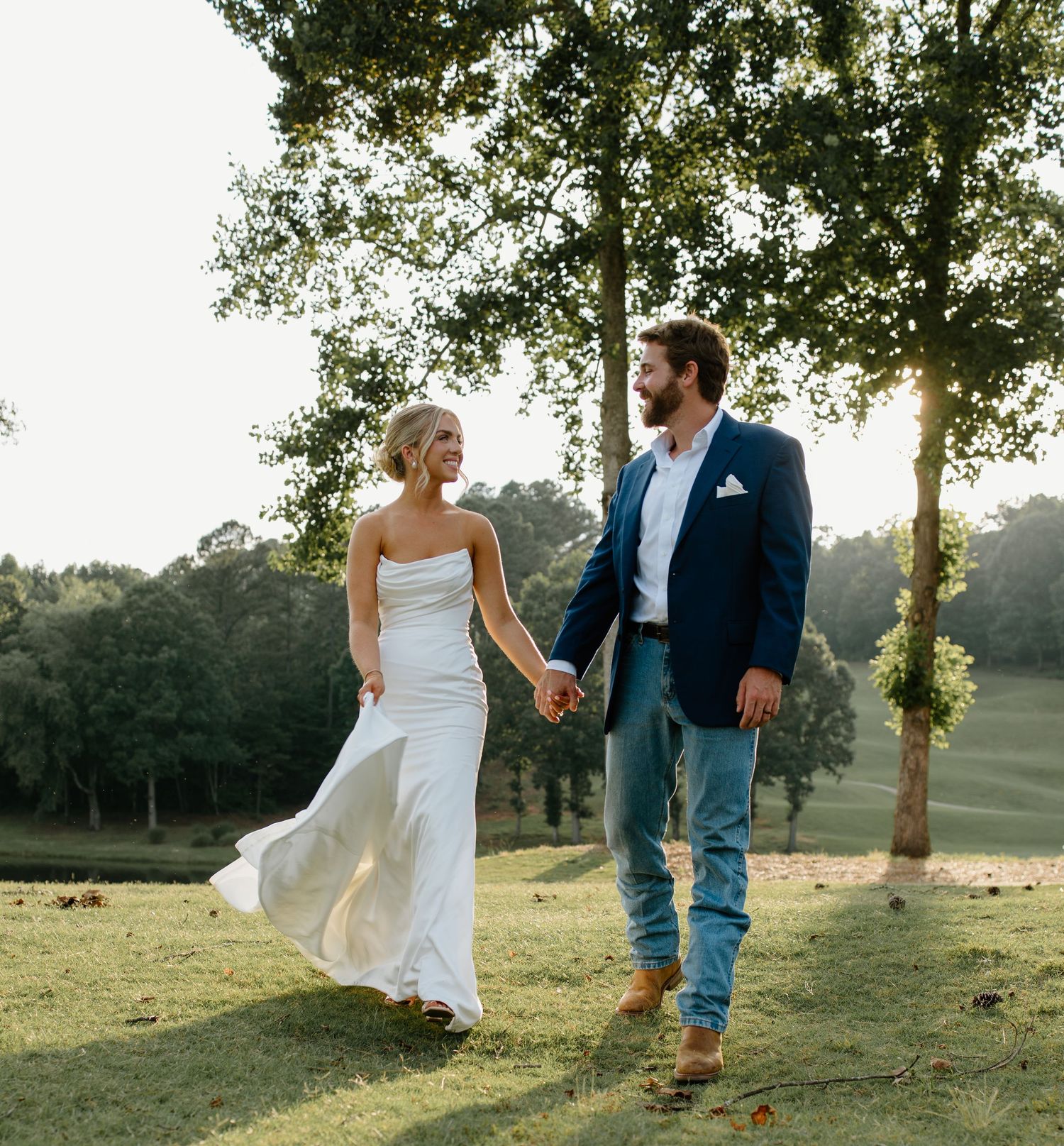 A couple walks hand in hand through a sunlit outdoor setting in wedding attire.