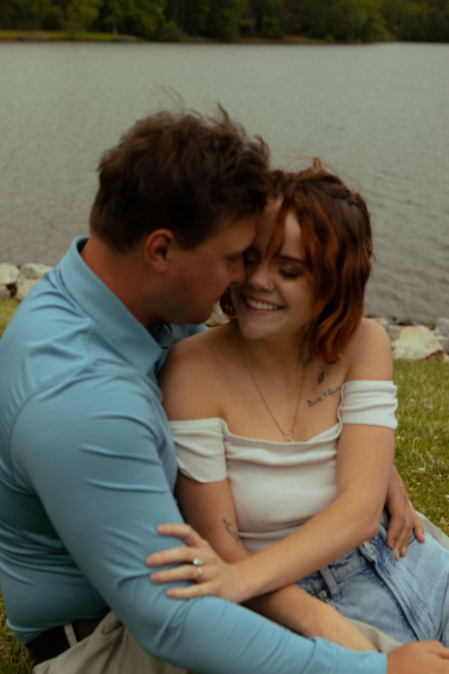 A romantic embrace captured between a couple on grass near a waterfront.