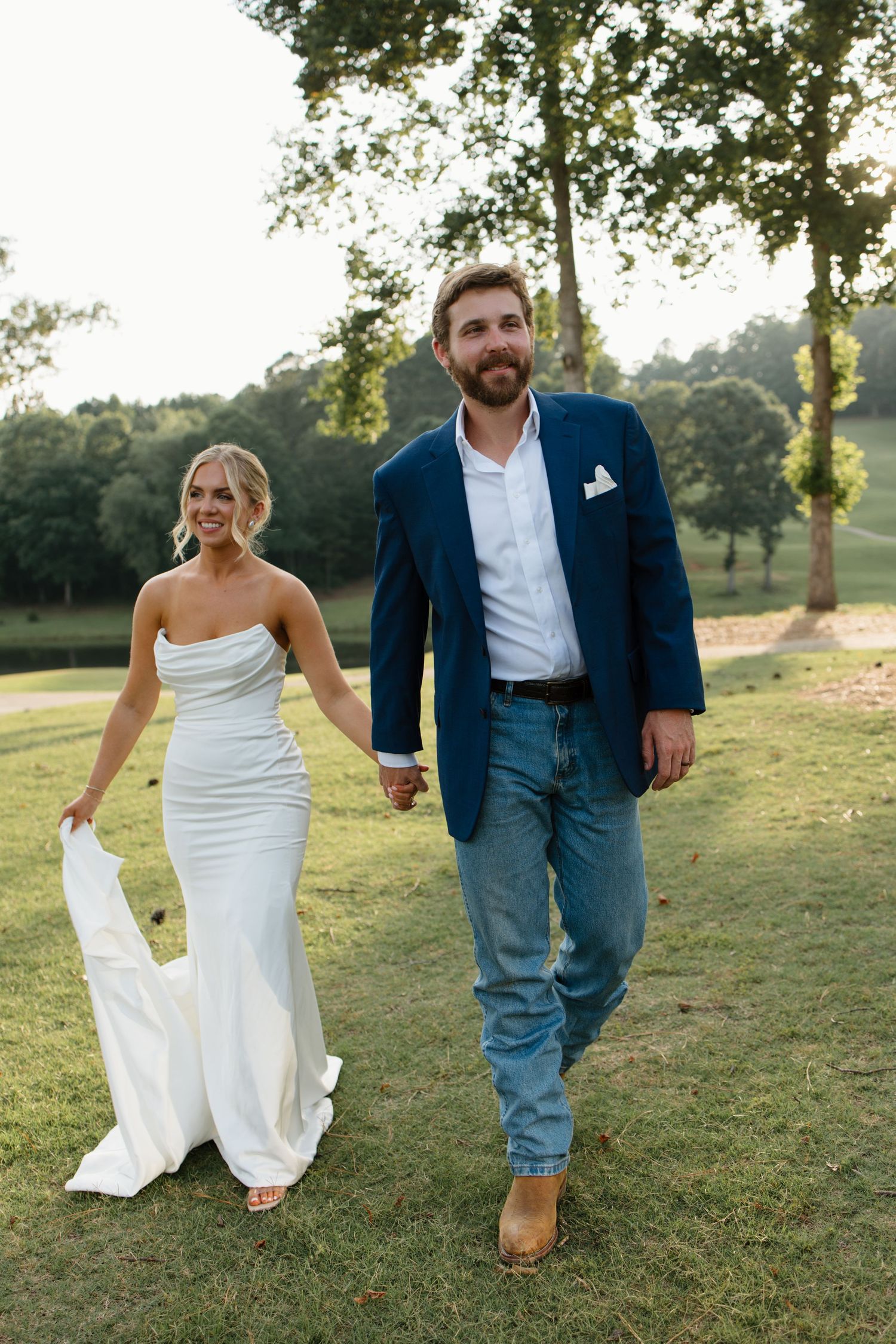 A bride and groom walk and laugh together on a grassy lawn during their wedding photo session at sunset.