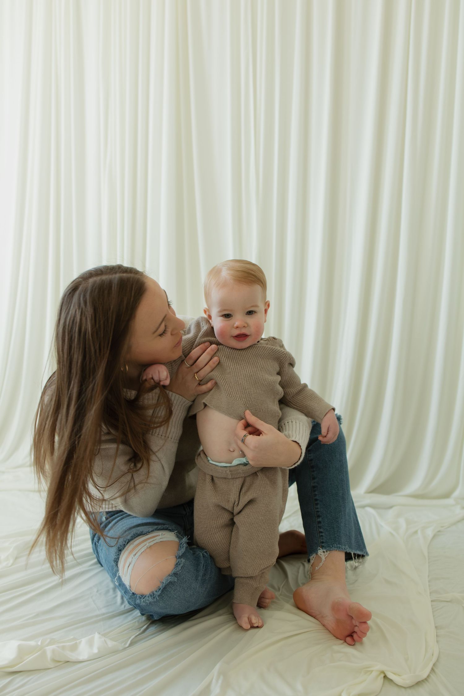 A tender moment between parent and baby sitting together against a white curtain backdrop.