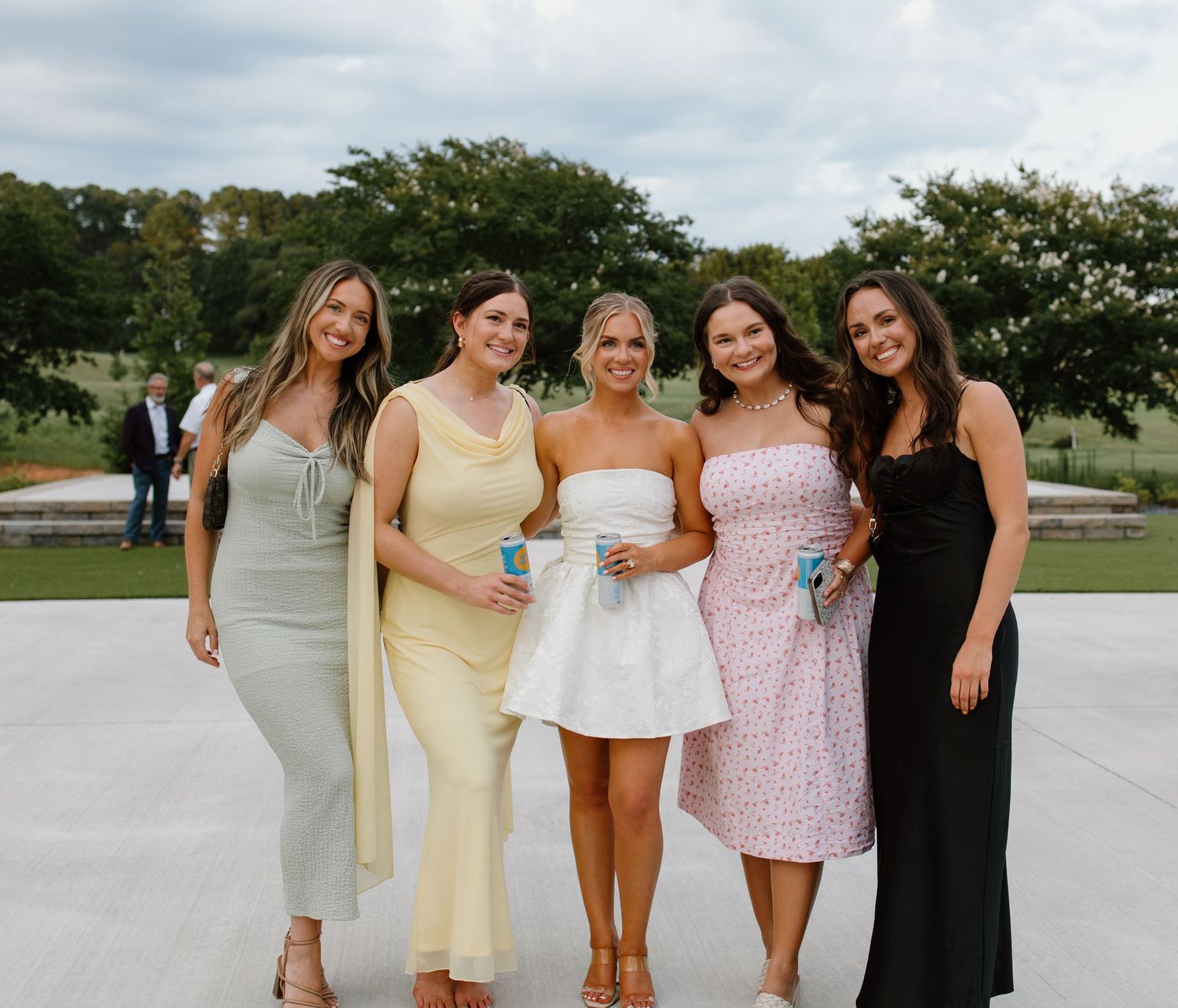 Five women in elegant dresses pose together on a concrete patio with trees and greenery in the background.